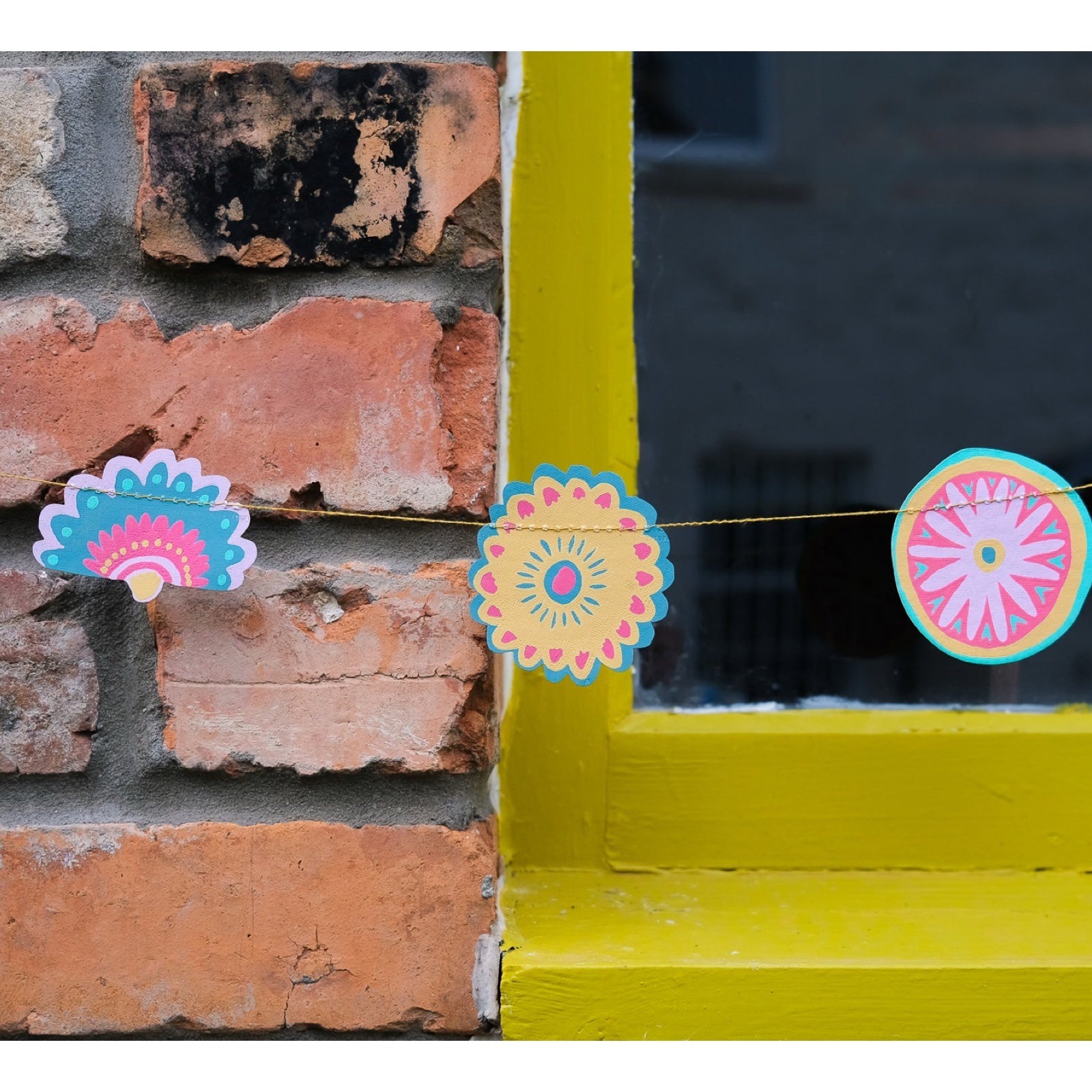 Folk-inspired garland, featuring twenty screen-printed decorations influenced by botanical illustrations, folk patterns, and symbology in summer shades of pink, yellow, turquoise and purple hanging against a brick wall.