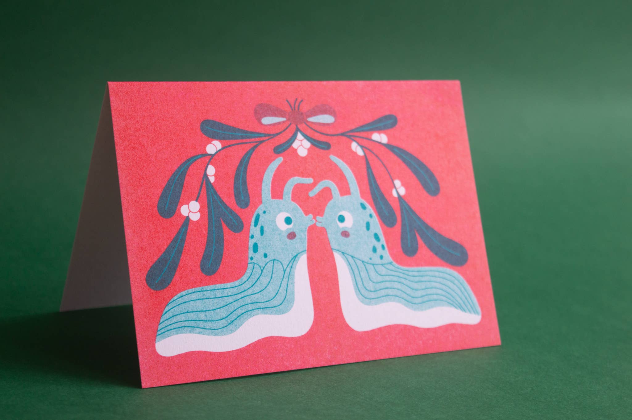 A greeting card standing upright on a dark green background. The card features a whimsical illustration of two blue slugs facing each other under a red bow and dark blue mistletoe.