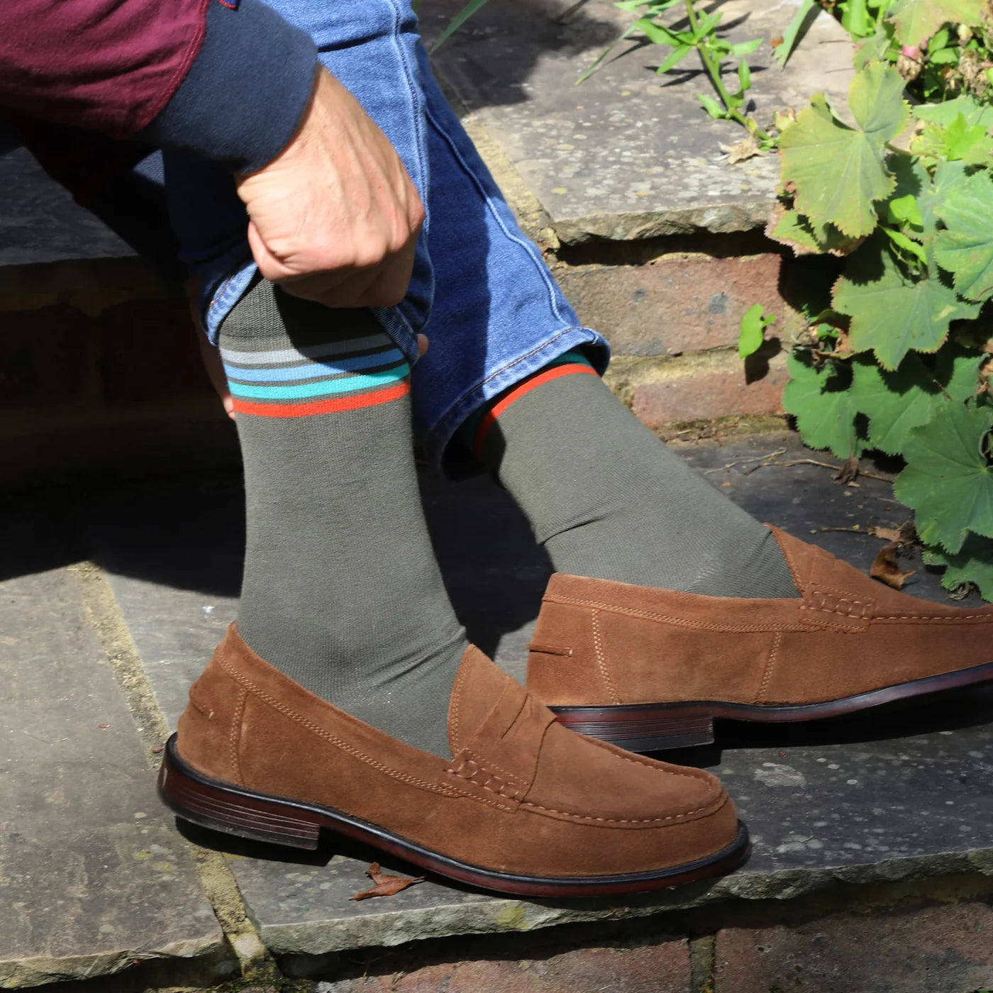 A person sitting outdoors on stone steps wearing brown suede penny loafers, olive green crew socks with colourful stripes near the top, and blue jeans.