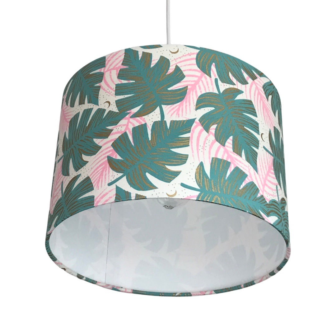 Close up of a drum lampshade covered in white fabric with green monstera leaves, pink leaves and gold detail.
