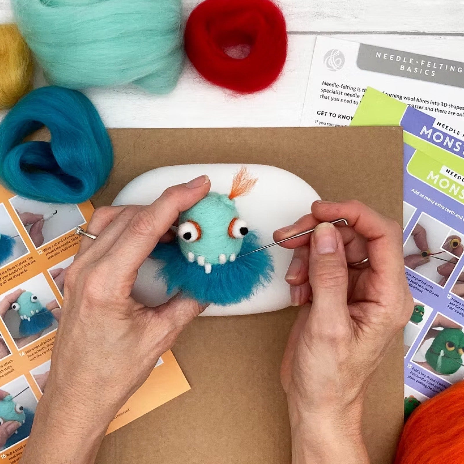 Person needle-felting a blue monster with colourful felt and craft materials on a table.