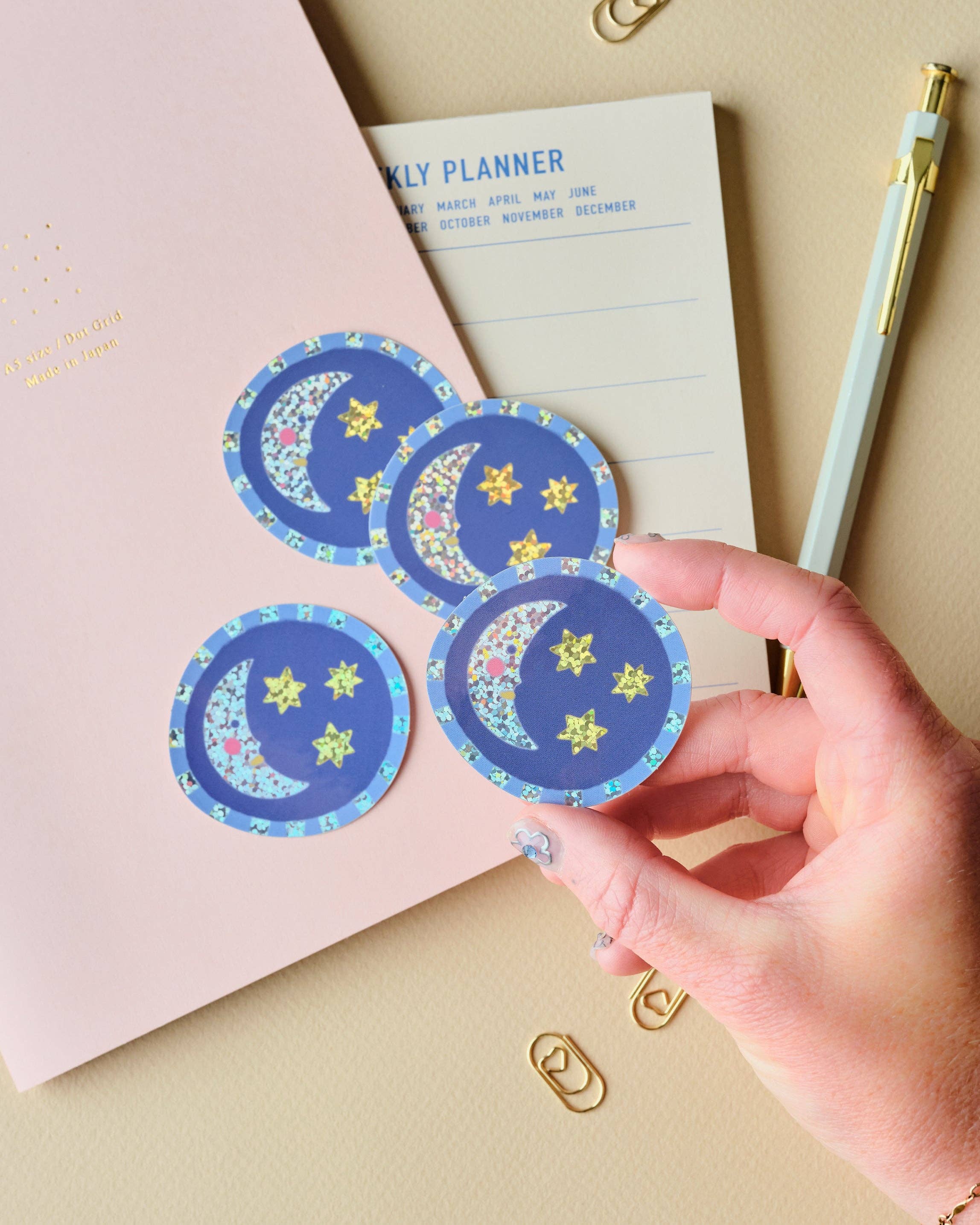 A hand holds three round blue stickers with a crescent moon and star pattern, placed over a pink notebook and a cream weekly planner on a beige desk with gold paperclips and a pen.