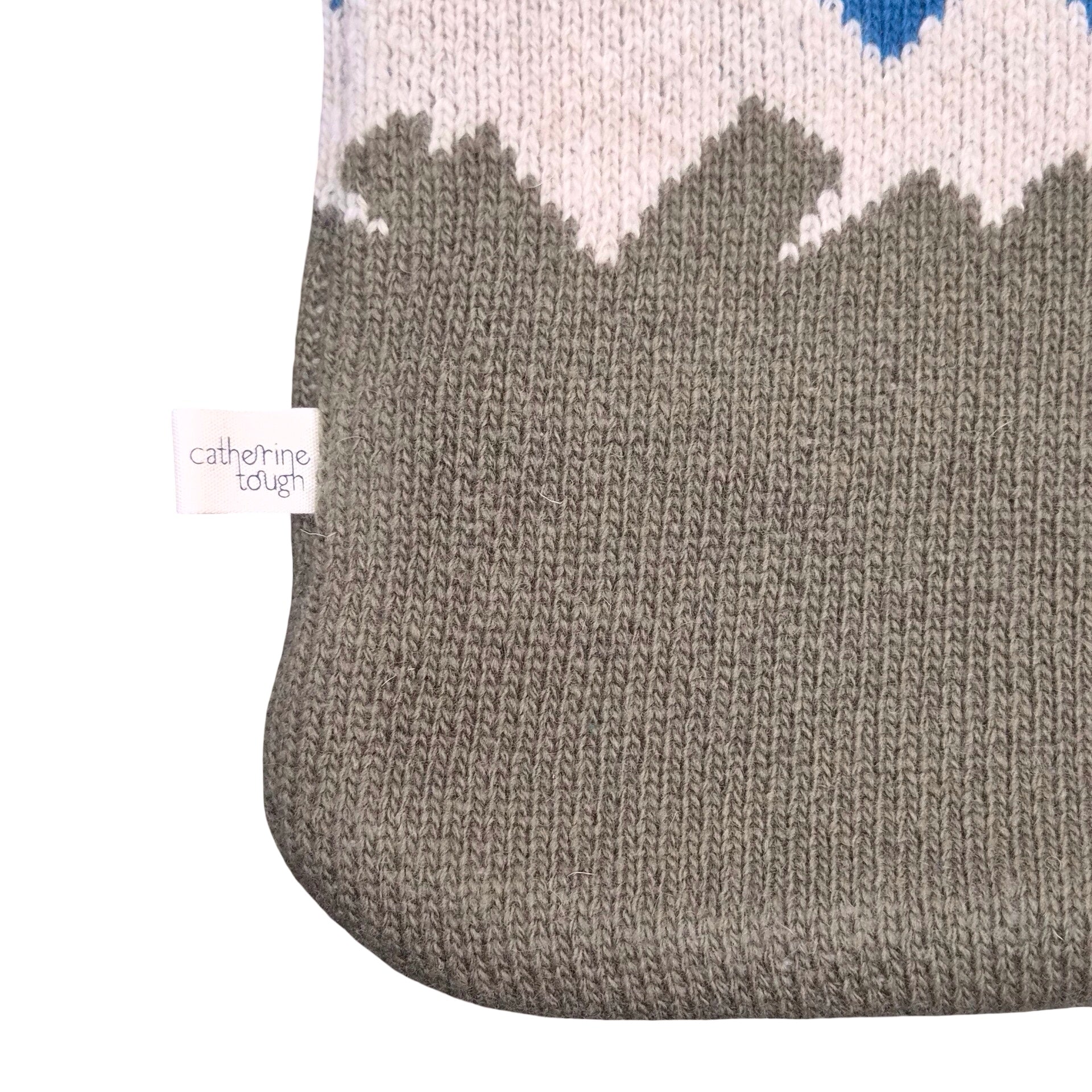 Close-up of a knitted water bottle showing a mountain pattern with a large grey section, a white section above, and a small blue accent at the top. A small fabric tag reads 'catherine tough'.