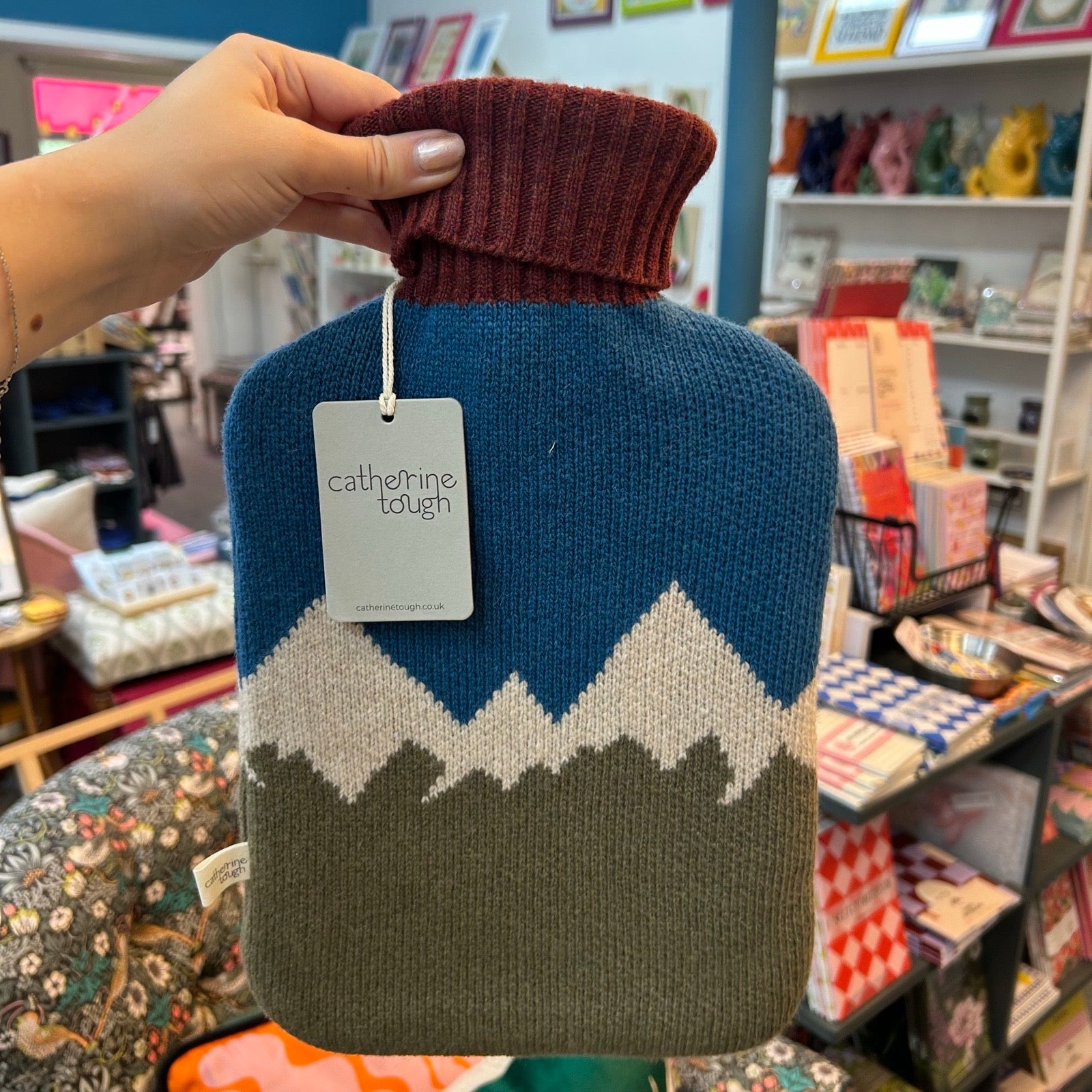 A hand holds a knitted hot water bottle cover with a mountain pattern. The cover has a dark blue top section, an off-white middle section, and a muted olive-brown bottom section, with a dark burgundy turtleneck collar.