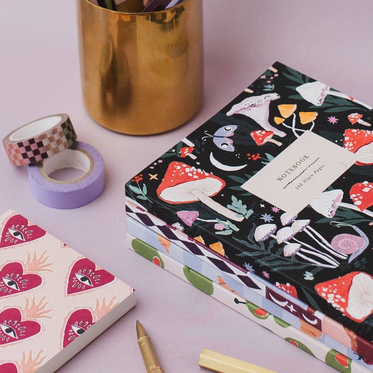 A stack of illustrated notebooks with a dark forest-themed cover featuring red mushrooms, styled on a pink surface with washi tape, a gold cup, and a pen.