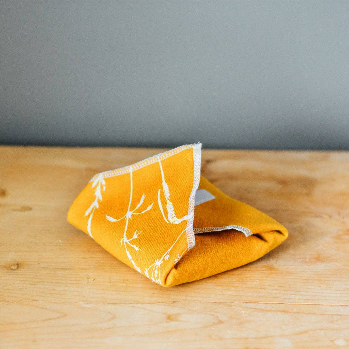 A folded mustard yellow fabric cloth with a white botanical line pattern and white stitched edges, resting on a light wood tabletop against a grey wall.