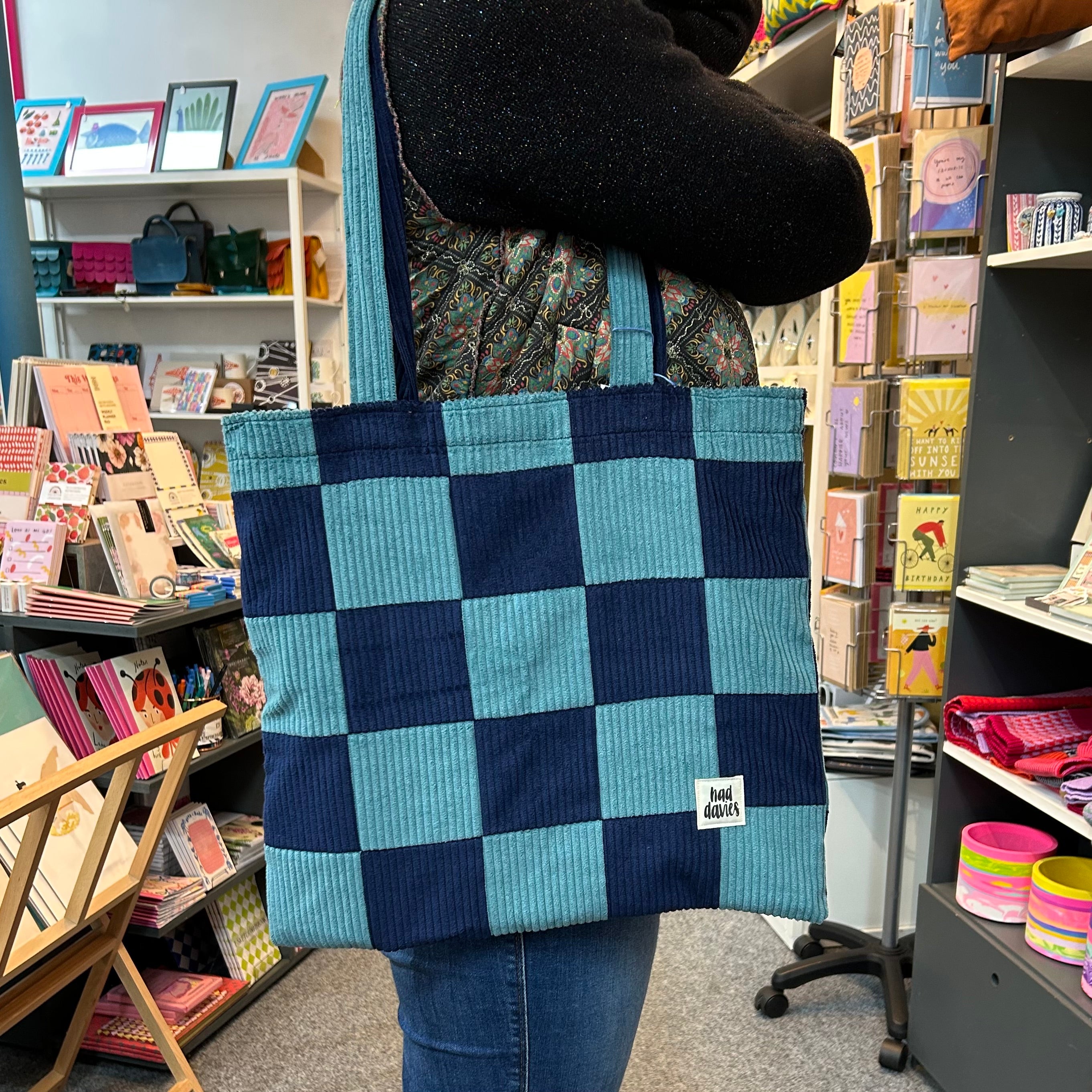 Person holding a blue and navy checkered tote bag in a bookstore setting.