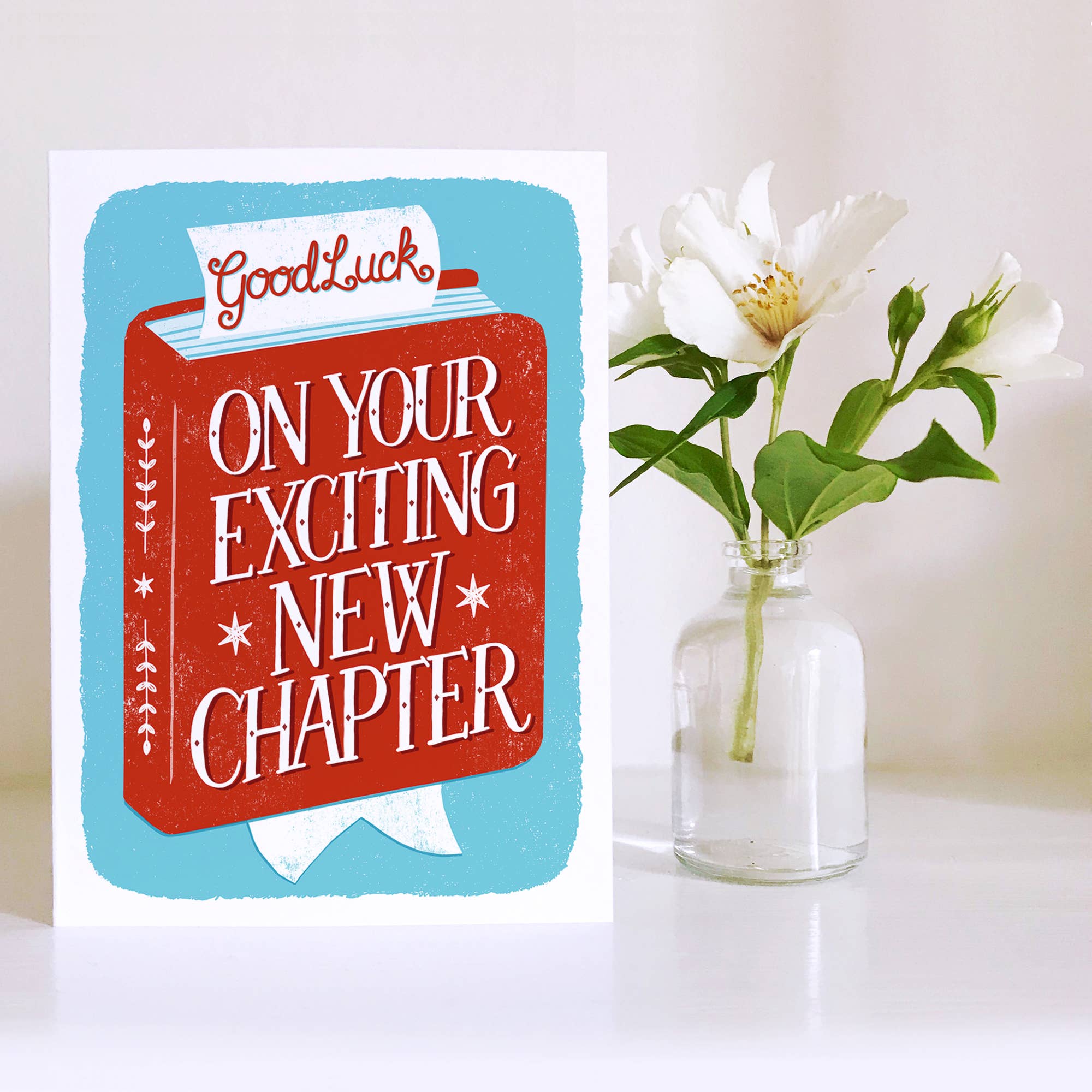 Greeting card with 'Good Luck on Your Exciting New Chapter' text next to a vase with white flowers on a light background.