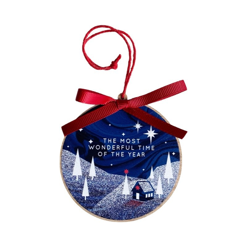 Circular Christmas decoration featuring an illustration of a wooden lodge in a forest under the northern lights and the text 'the most wonderful time of the year'.