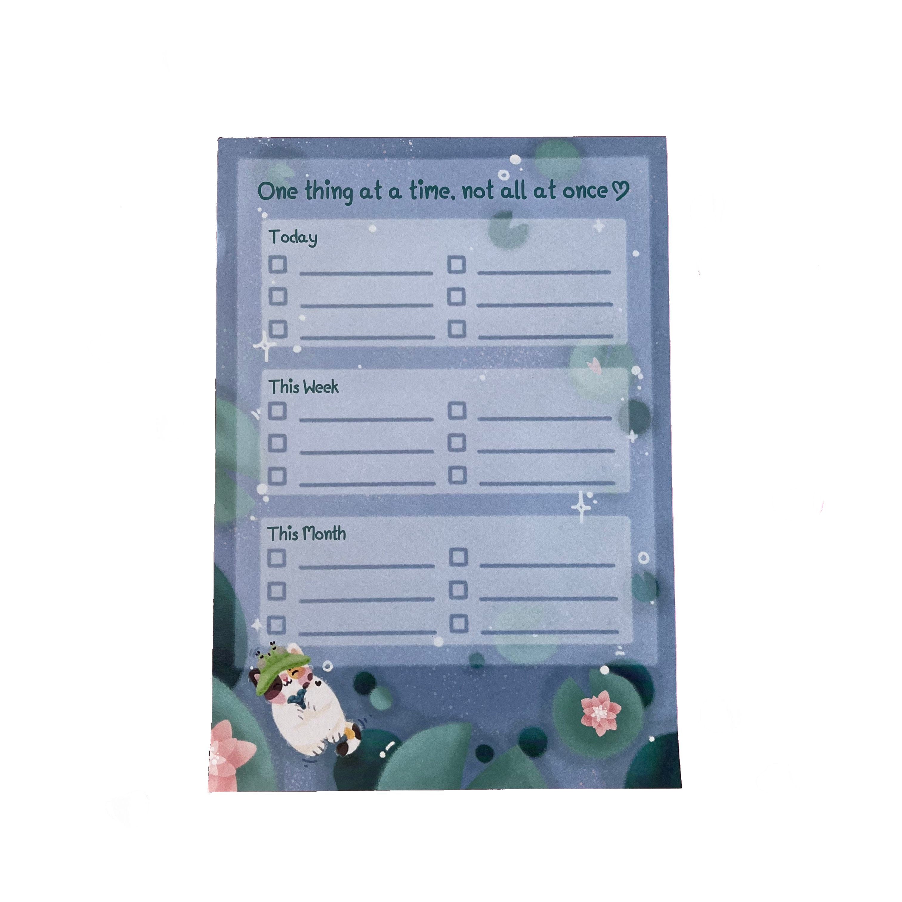 A 50-page tear-off lined notepad to help with your to do list items. With three sections to help prioritise tasks, labelled: 'today', 'this week', and 'this month', with check boxes. Text at top reads 'one thing at a time, not all at once'. The background features waterlily pads and a calico cat swimming on it's back while holding a heart shaped rock. 