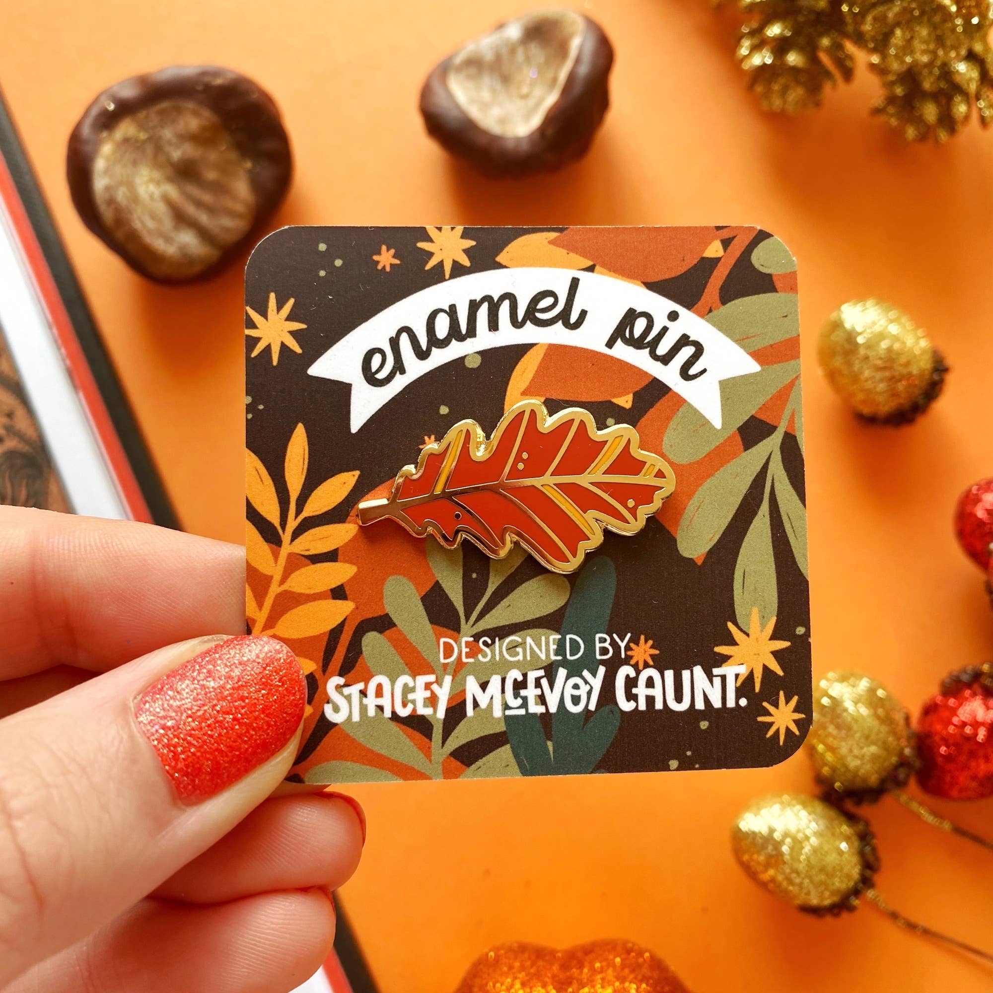 An autumn-themed enamel pin shaped like an oak leaf, held by a hand on its backing card. The pin features a warm orange and red leaf with gold-toned metal outlines, set against an orange background with colourful chestnuts.