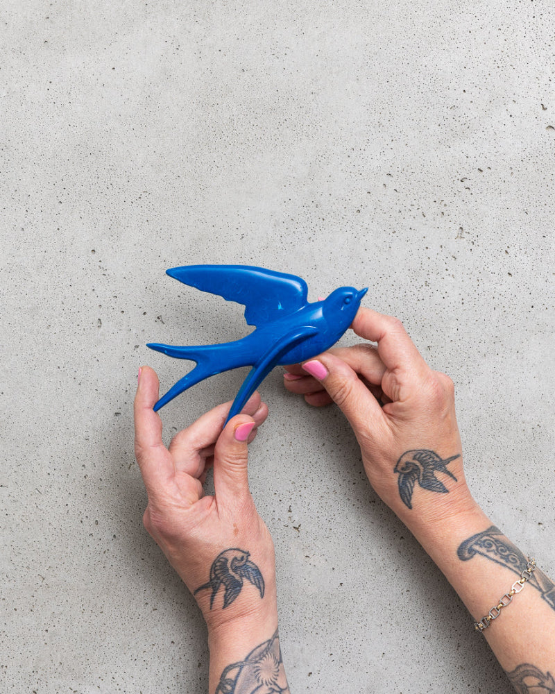 Blue bird-shaped object held by hands with tattoos on a grey surface.