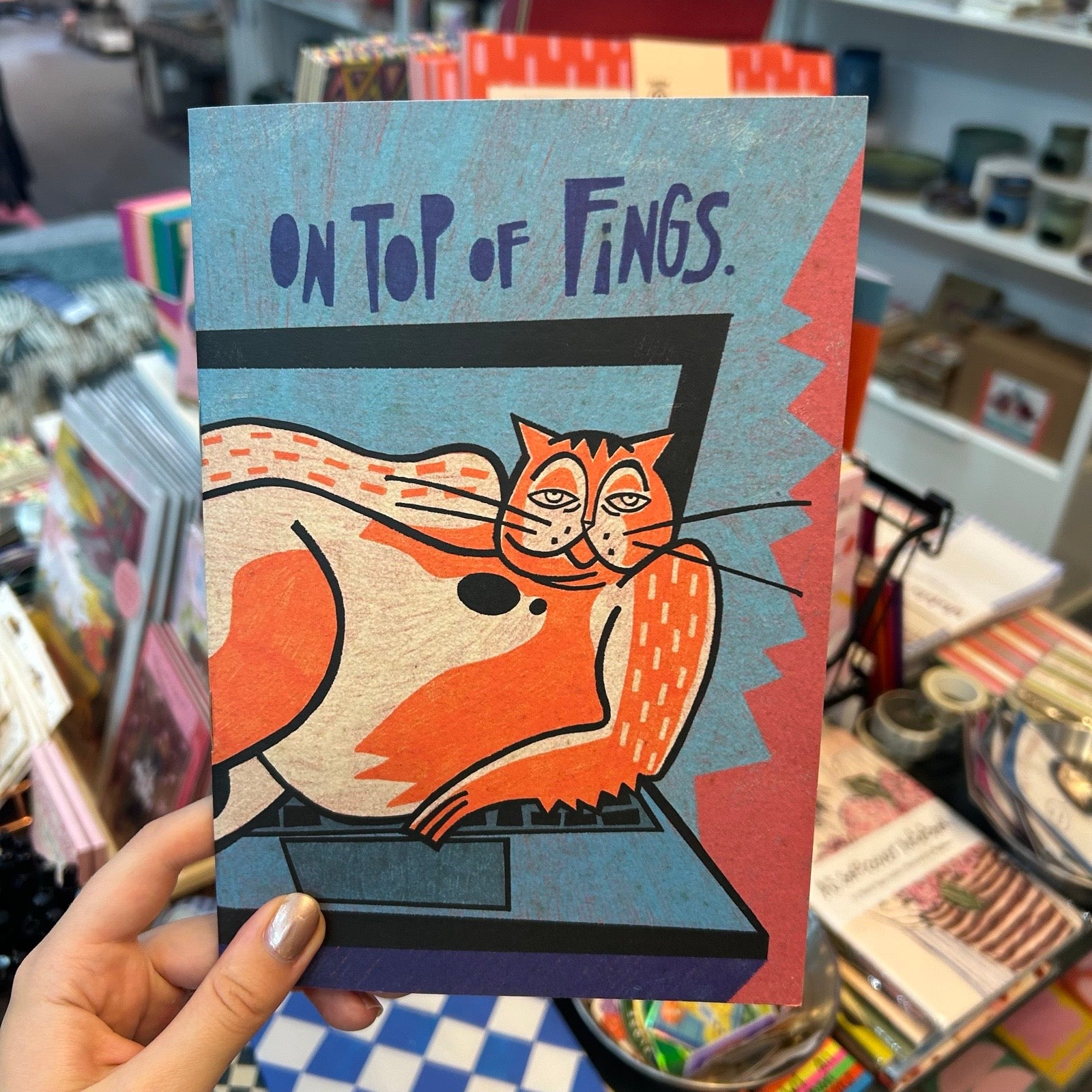 A hand holds a notebook featuring a stylised orange cat sleeping on a laptop with the text 'ON TOP OF FINGS.' The background shows a busy gift shop or stationery store interior with shelves and other items.