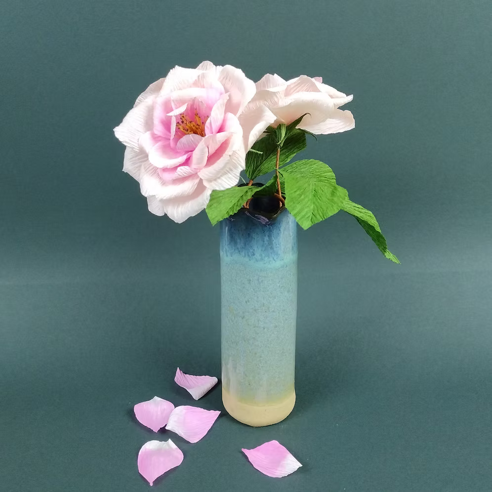 Vase with pink flowers and petals on a teal background.