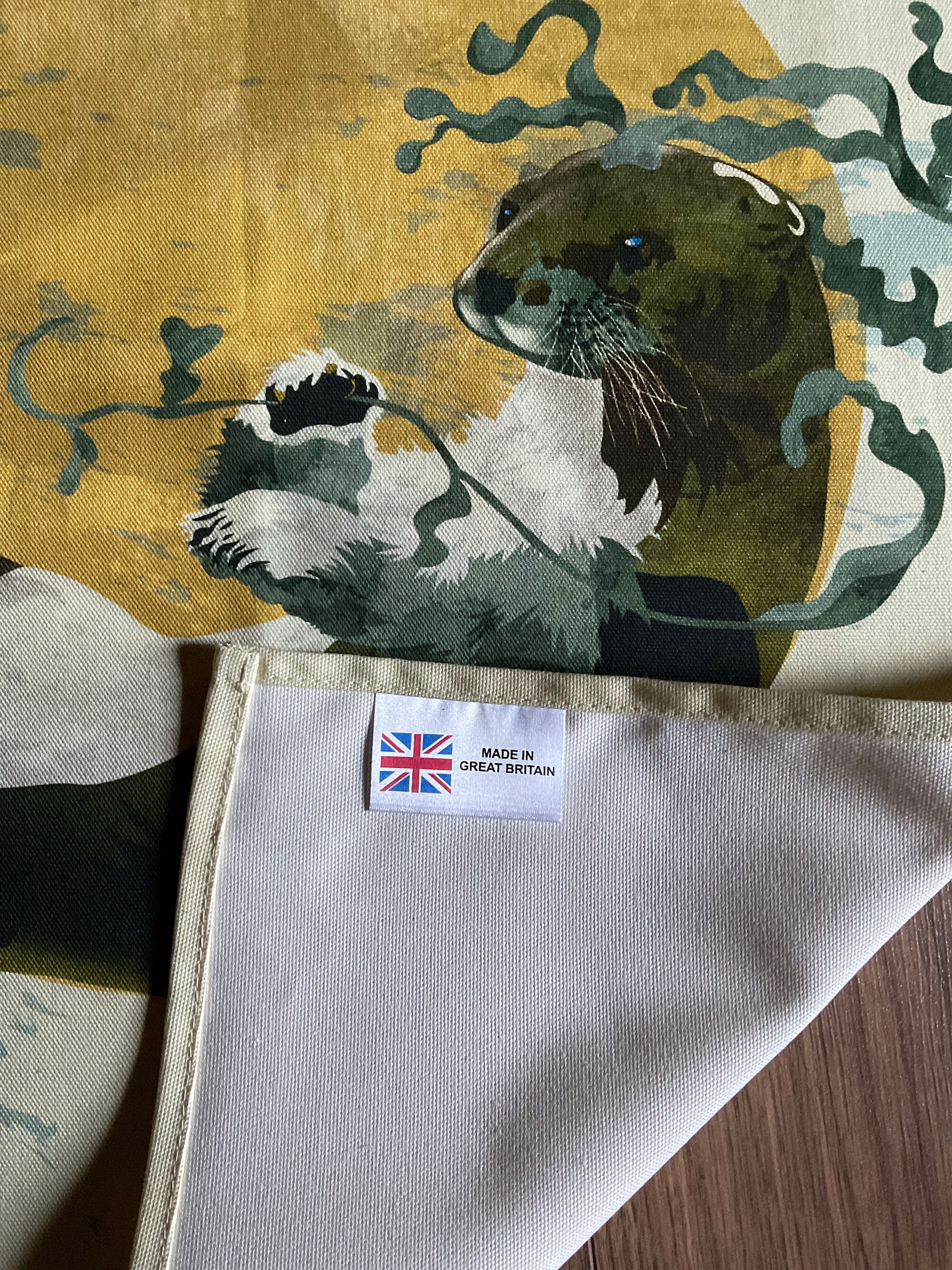 A tea towel featuring an illustration of an otter, in a mustard circle against an off-white background.