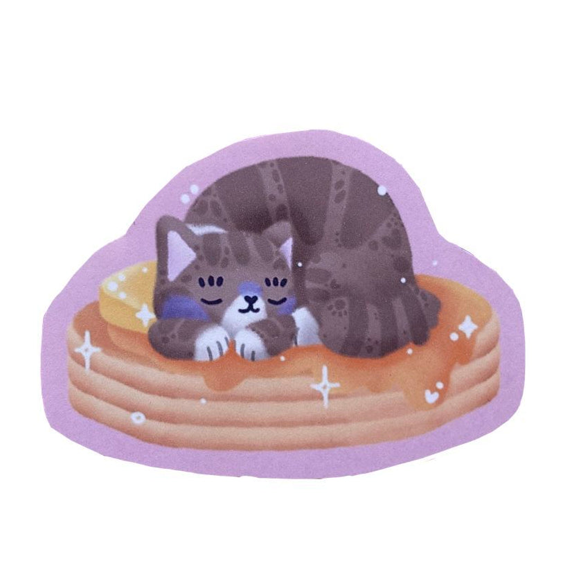 Handmade sticker of a brown and white tabby cat, sleeping on a stack of pancakes with butter and syrup.