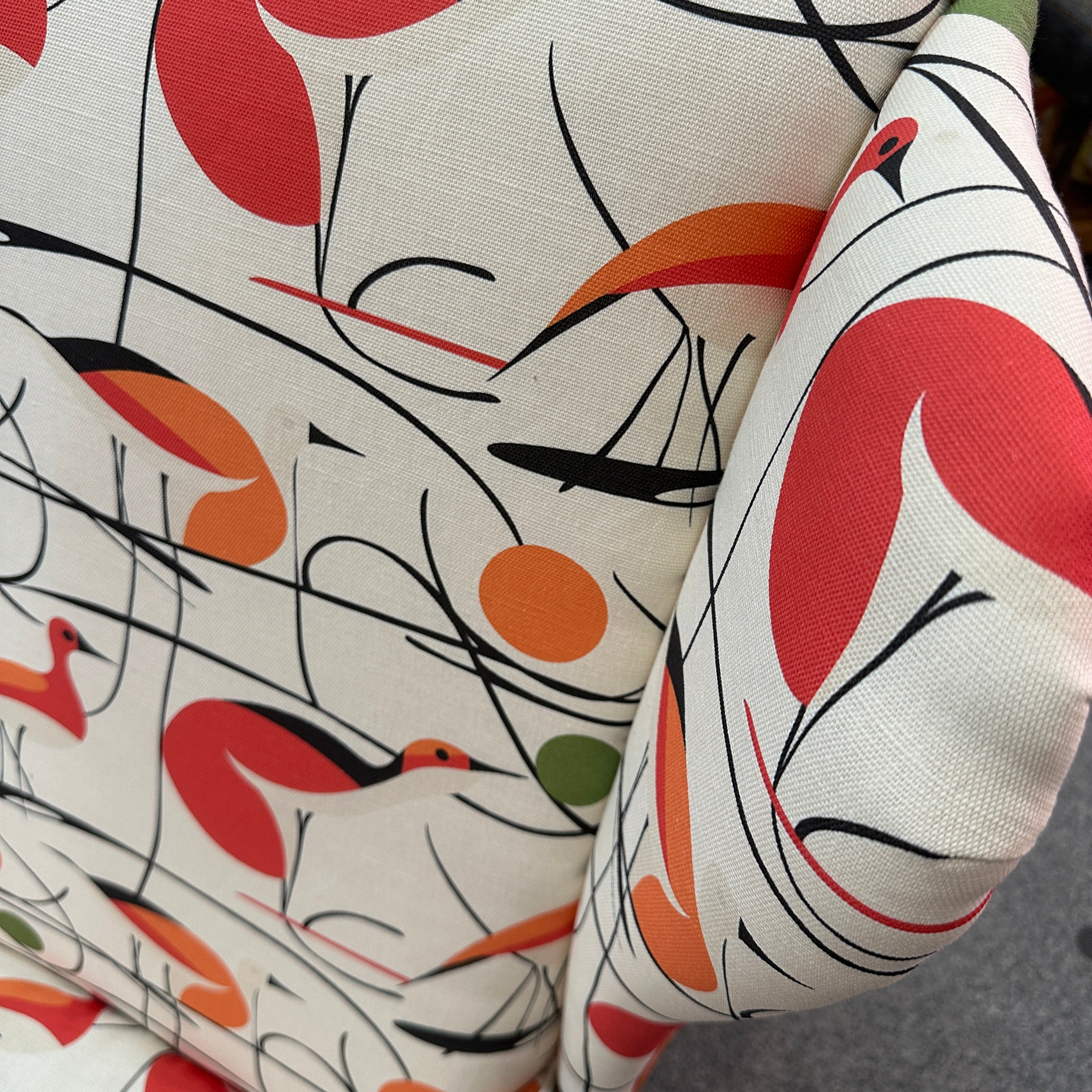 A close-up of a fabric surface with an abstract floral or botanical print. The pattern features large coral-red and orange leaf-like shapes, thin black curved lines, and small green accents on an off-white woven background.
