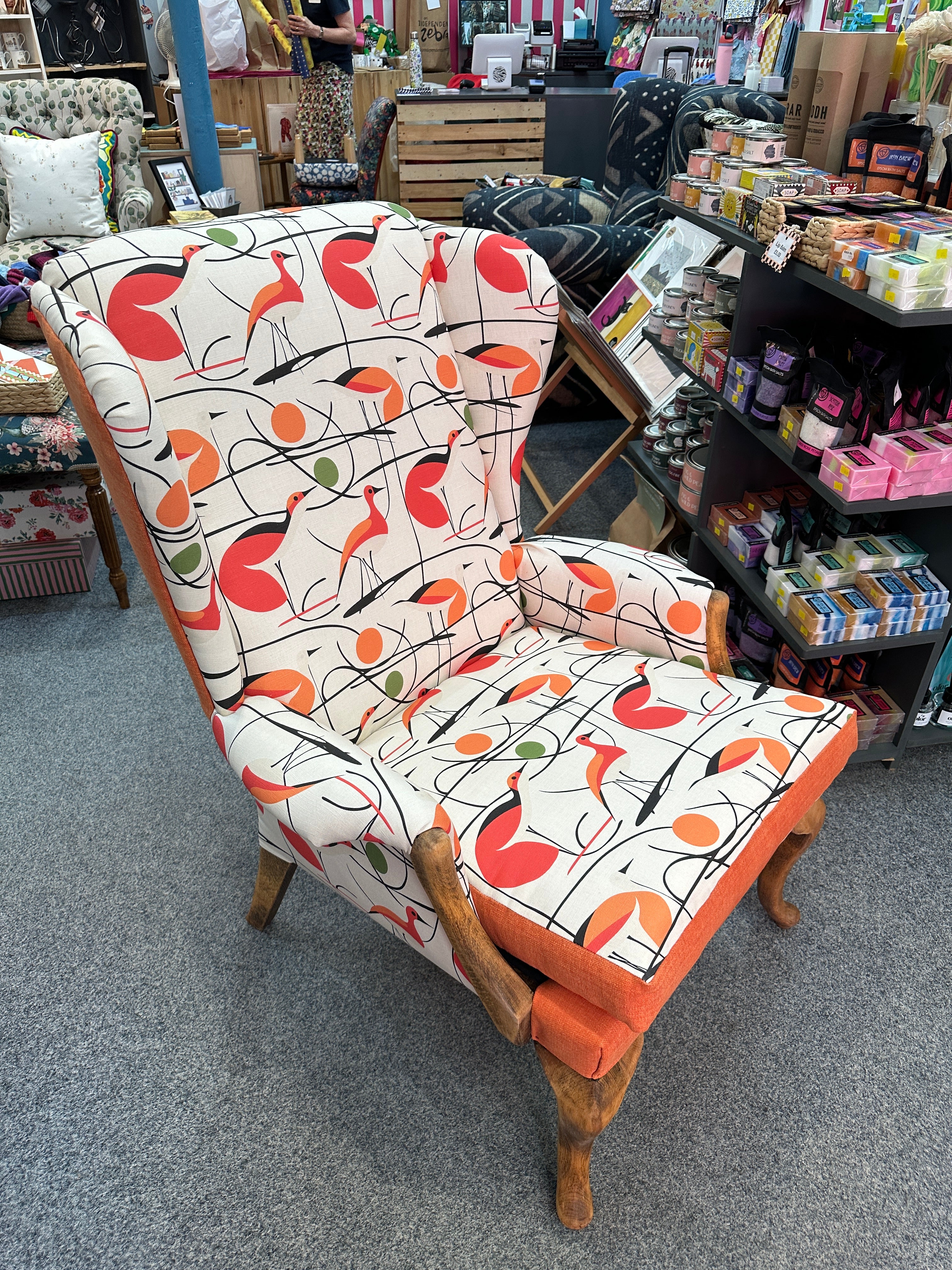 A mid-century modern accent chair with a cream fabric upholstered in a bold orange, red, and black abstract pattern and wooden legs, photographed in a furniture store.