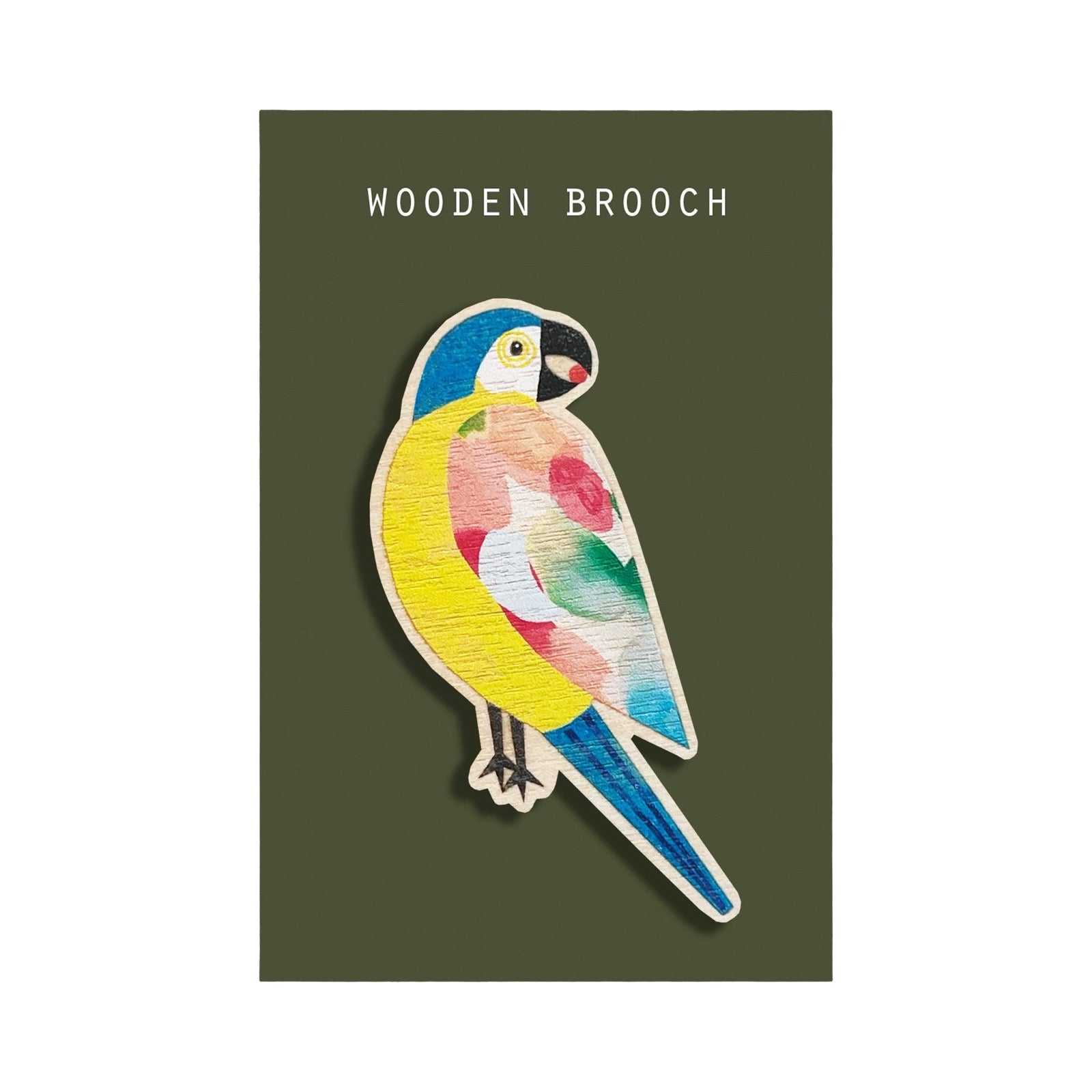 A colourful wooden bird brooch featuring a parrot design with yellow, blue, pink, and green tones, mounted on a dark olive green backing card with the text 'WOODEN BROOCH' at the top.