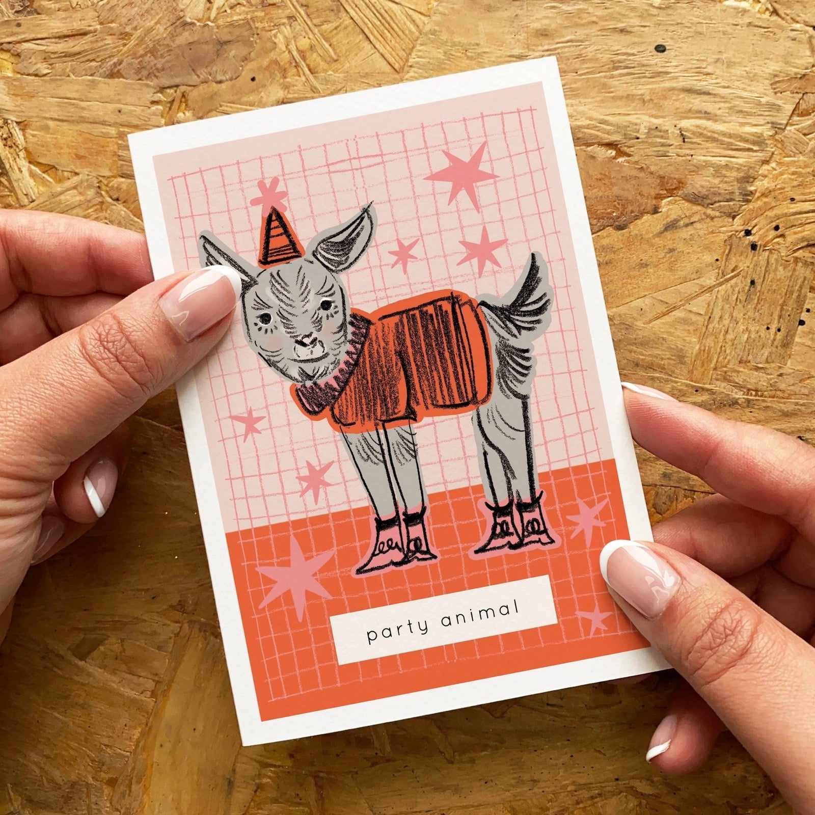 Card with a donkey illustration wearing a party hat and orange jumper, held by hands on a wooden surface.