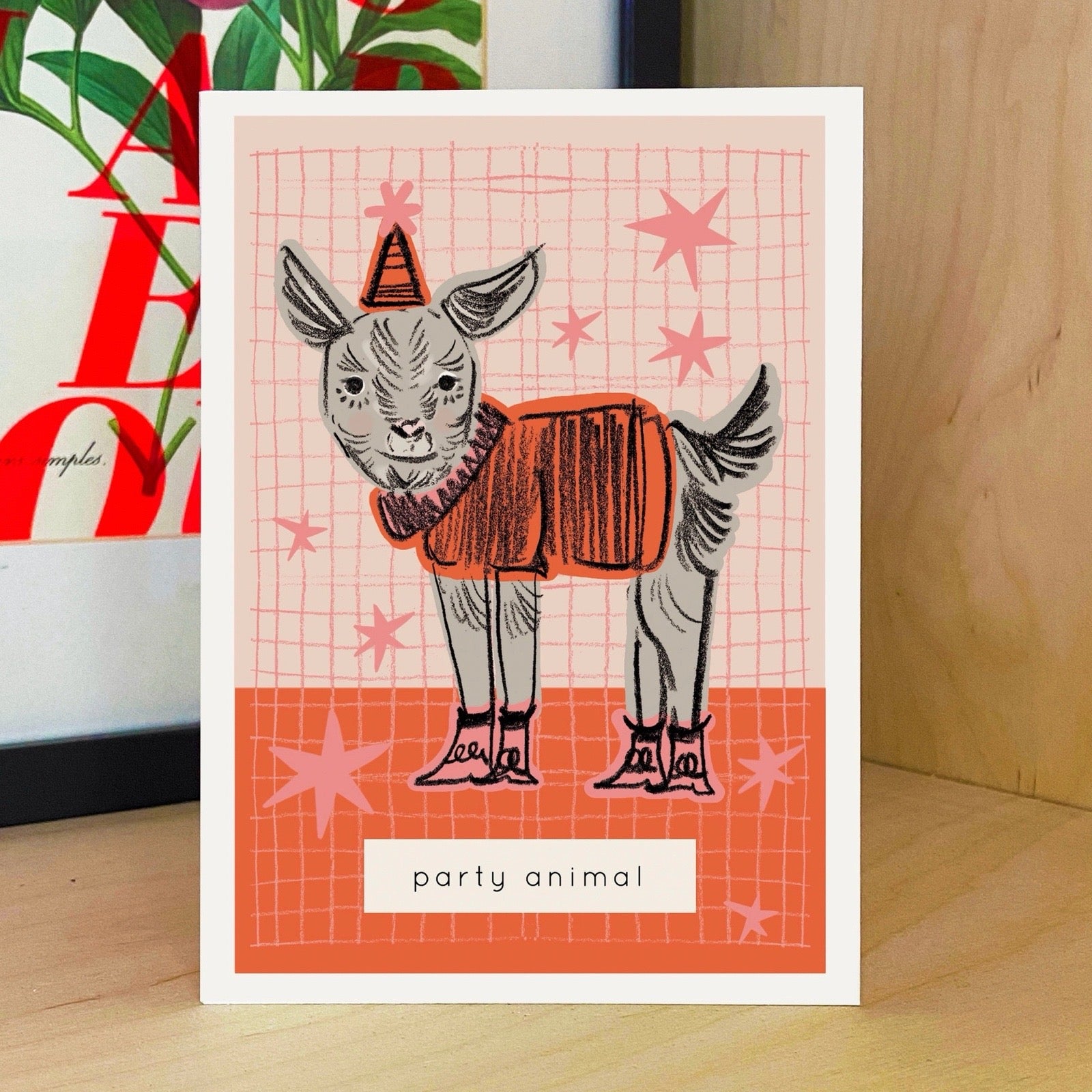 Card with illustration of a donkey wearing a party hat and sweater on a grid background.
