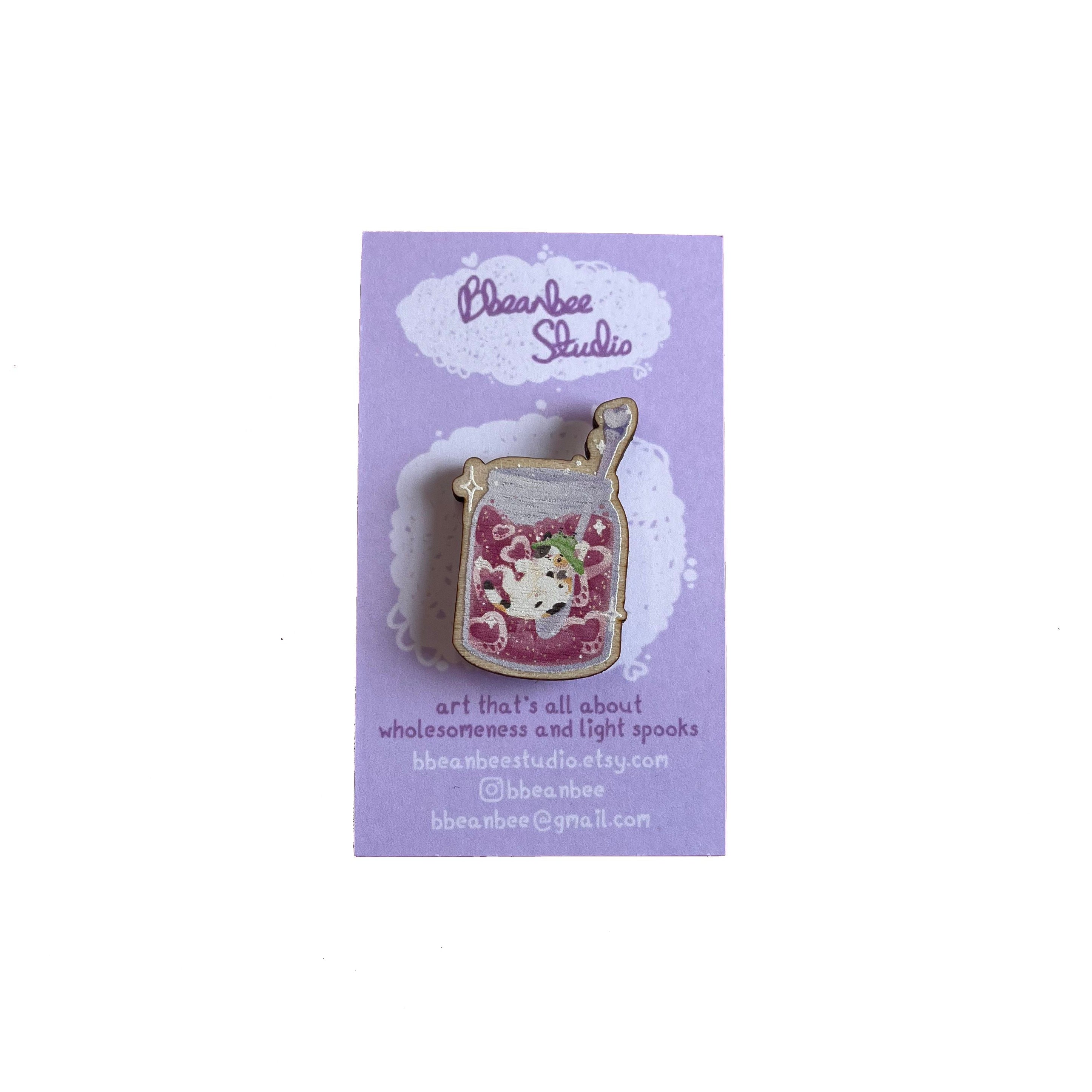 Wooden pin badge of a pawberry (heart-shaped strawberries with a paw shape inside) jam jar, with Bucket the calico cat inside. The spoon has a bee on it. This badge is printed onto 3mm thick responsibly sourced European birch plywood.