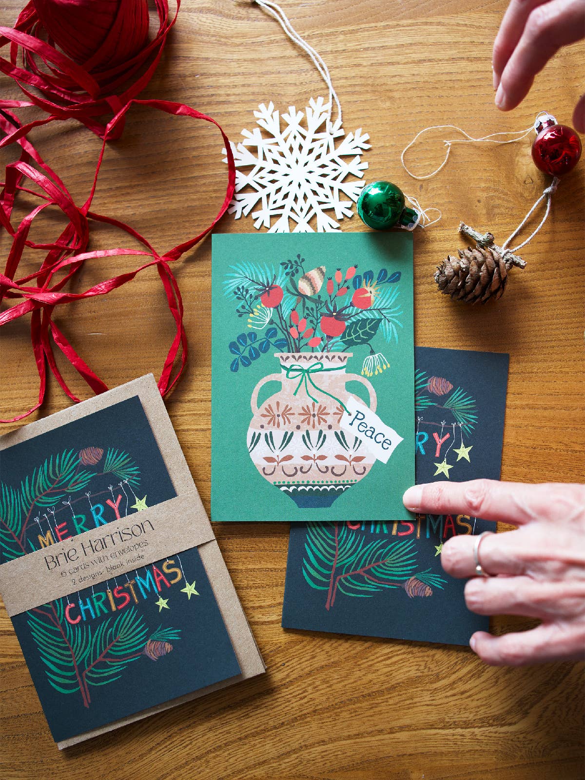 Christmas greeting cards on a wooden tabletop, with hands entering from the top and right to adjust the cards. The main product is a teal Christmas card featuring a white pitcher filled with red berries and green pine branches, with the word 'Peace' on the card.
