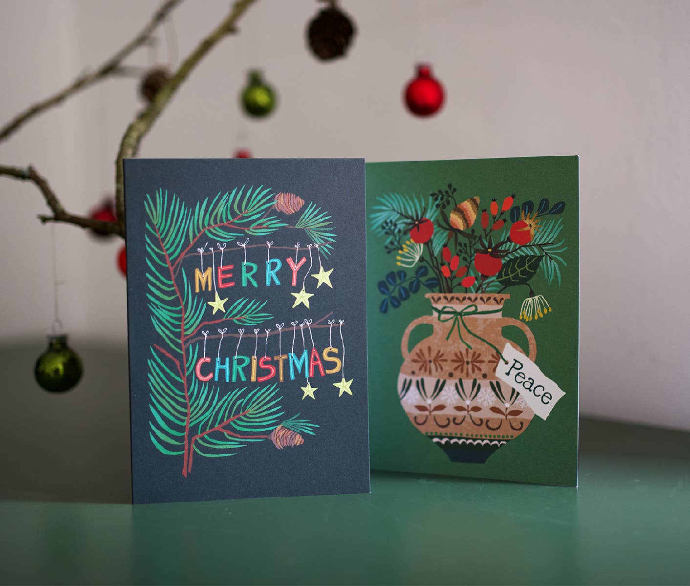 Two Christmas greeting cards displayed side by side on a teal surface against a light wall. The left card is dark blue with teal pine branches, colorful 'MERRY CHRISTMAS' lettering, and hanging lights.
