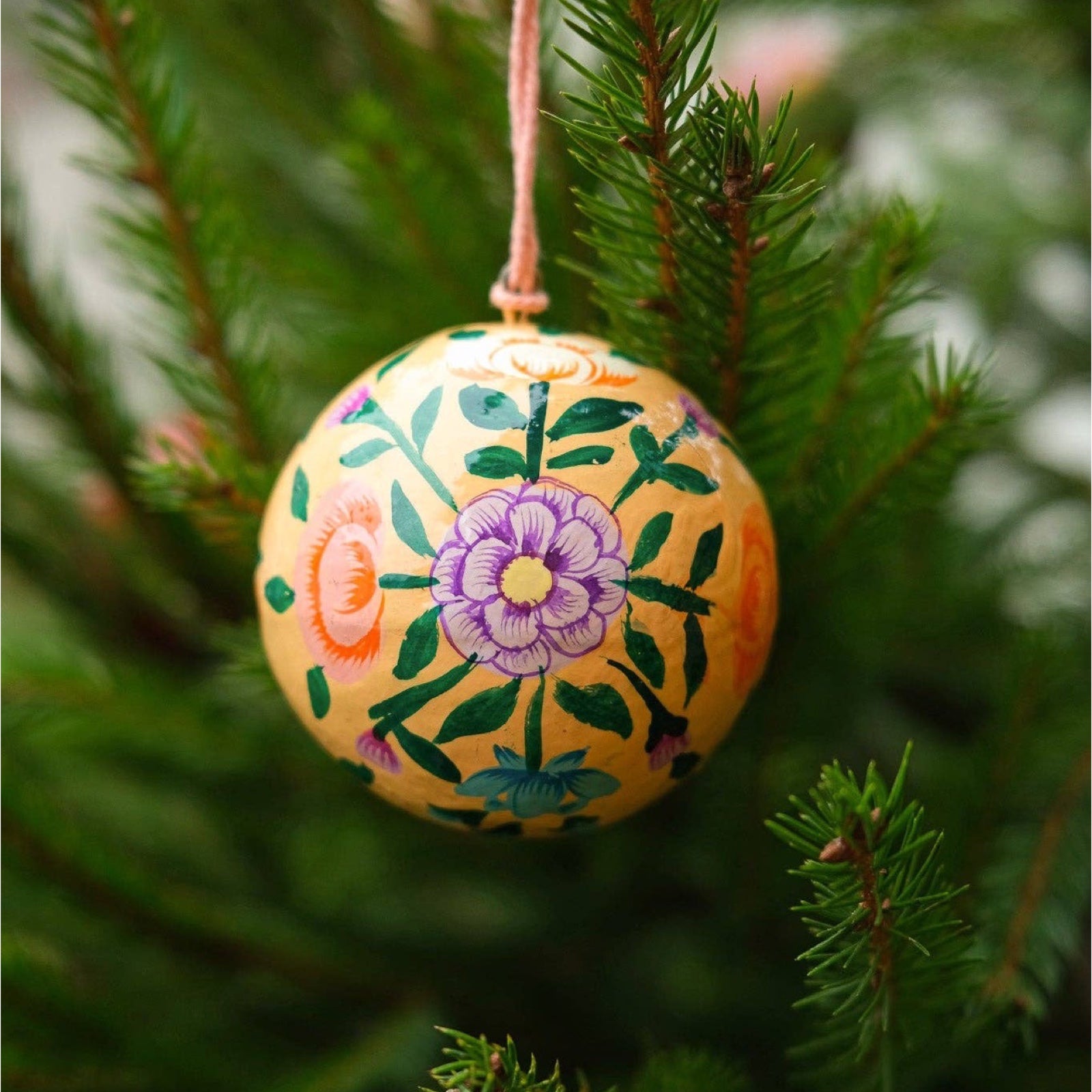 A round papier mache bauble in peach with a lilac, orange, green and turquoise floral design hanging on a Christmas tree.