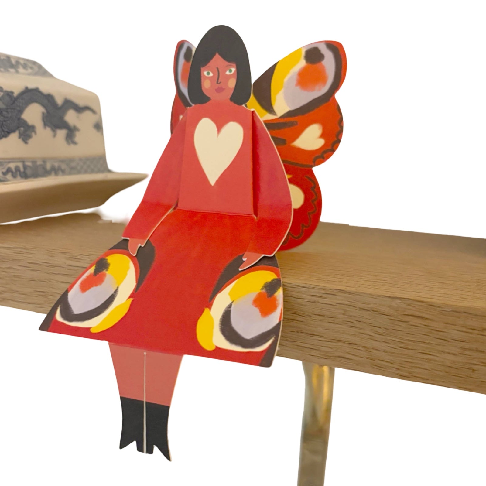 A butterfly woman card featuring a lady in a red dress with a white love heart and colourful wings that match the base of her dress sitting on a wooden shelf.