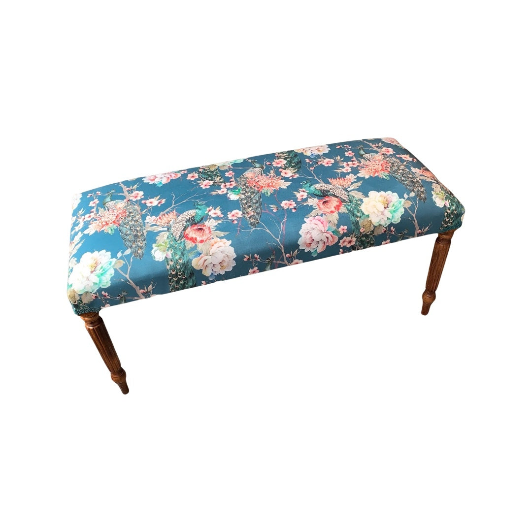 A hand-crafted rectangular bench covered in luxurious teal velvet with a floral pattern, featuring green peacocks and pink and white flowers, with teal binding and mahogany legs.
