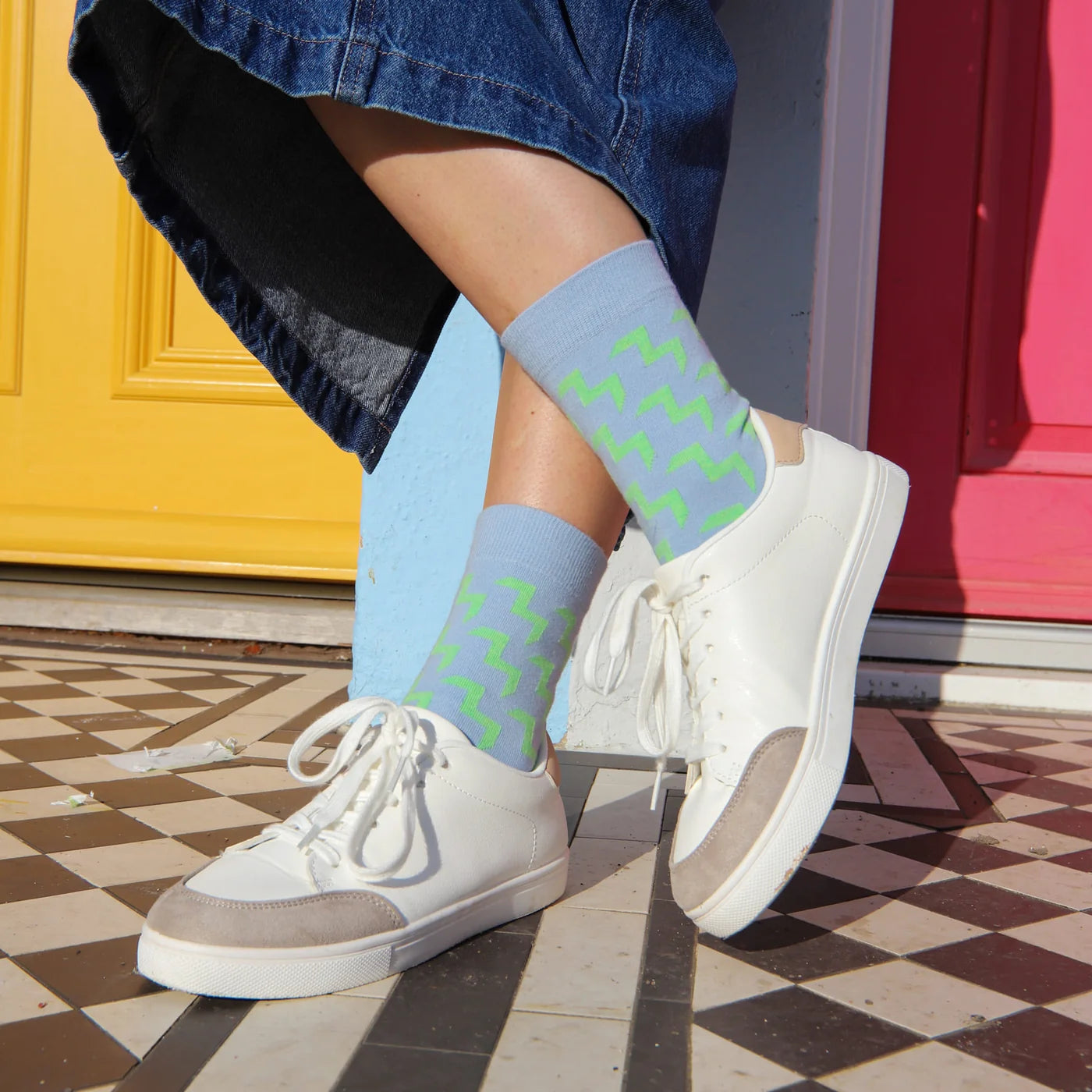 A person wearing light blue crew socks with a green abstract pattern, paired with white low-top sneakers and a blue denim skirt, standing on a checkered floor in front of a yellow and pink door.