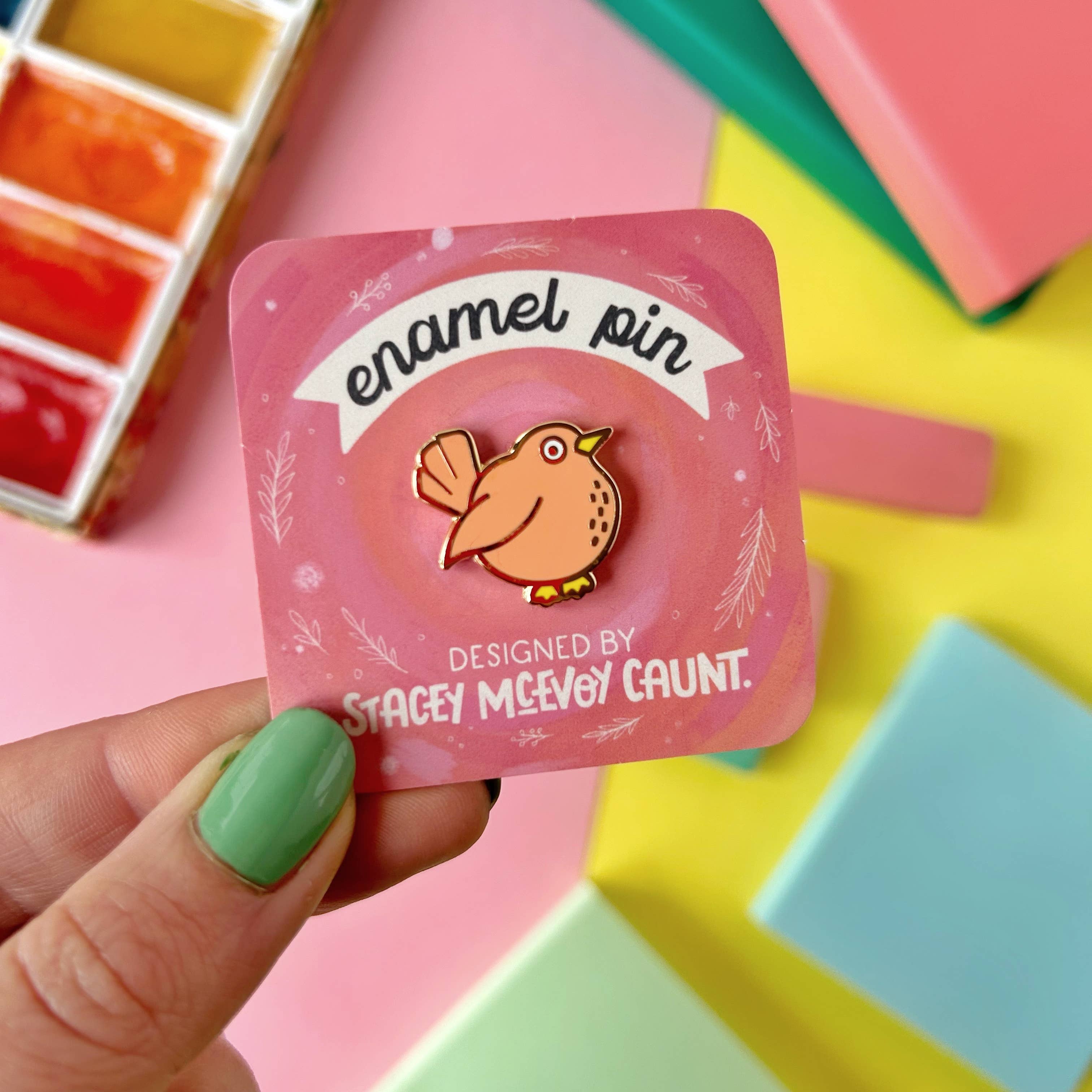 Enamel pin packaging with a bird design held by a hand against a colourful background.