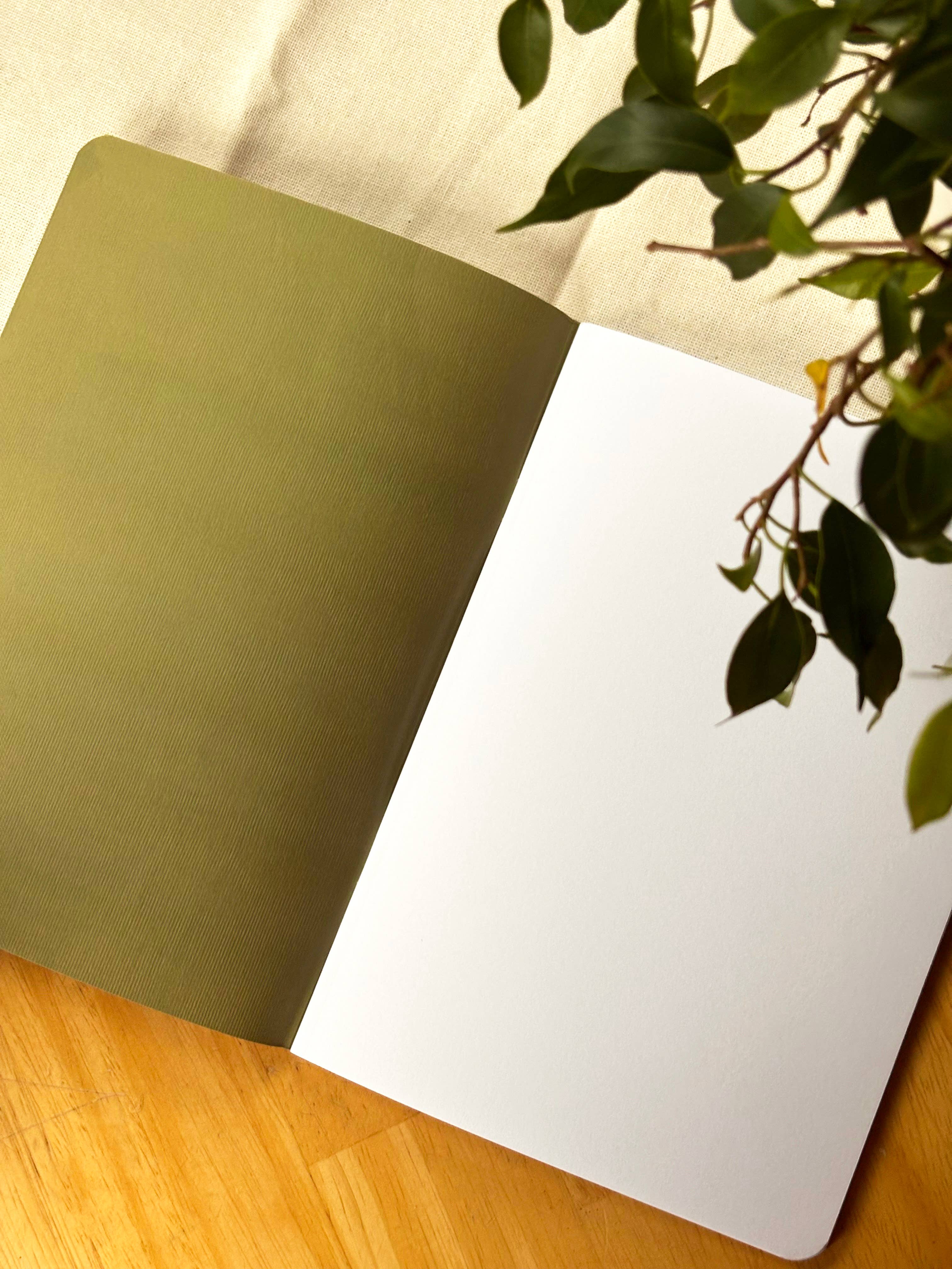 An open notebook with a muted olive green left page and blank white right page, photographed on a light wood surface with a beige fabric background and green plant leaves hanging in from the top right.