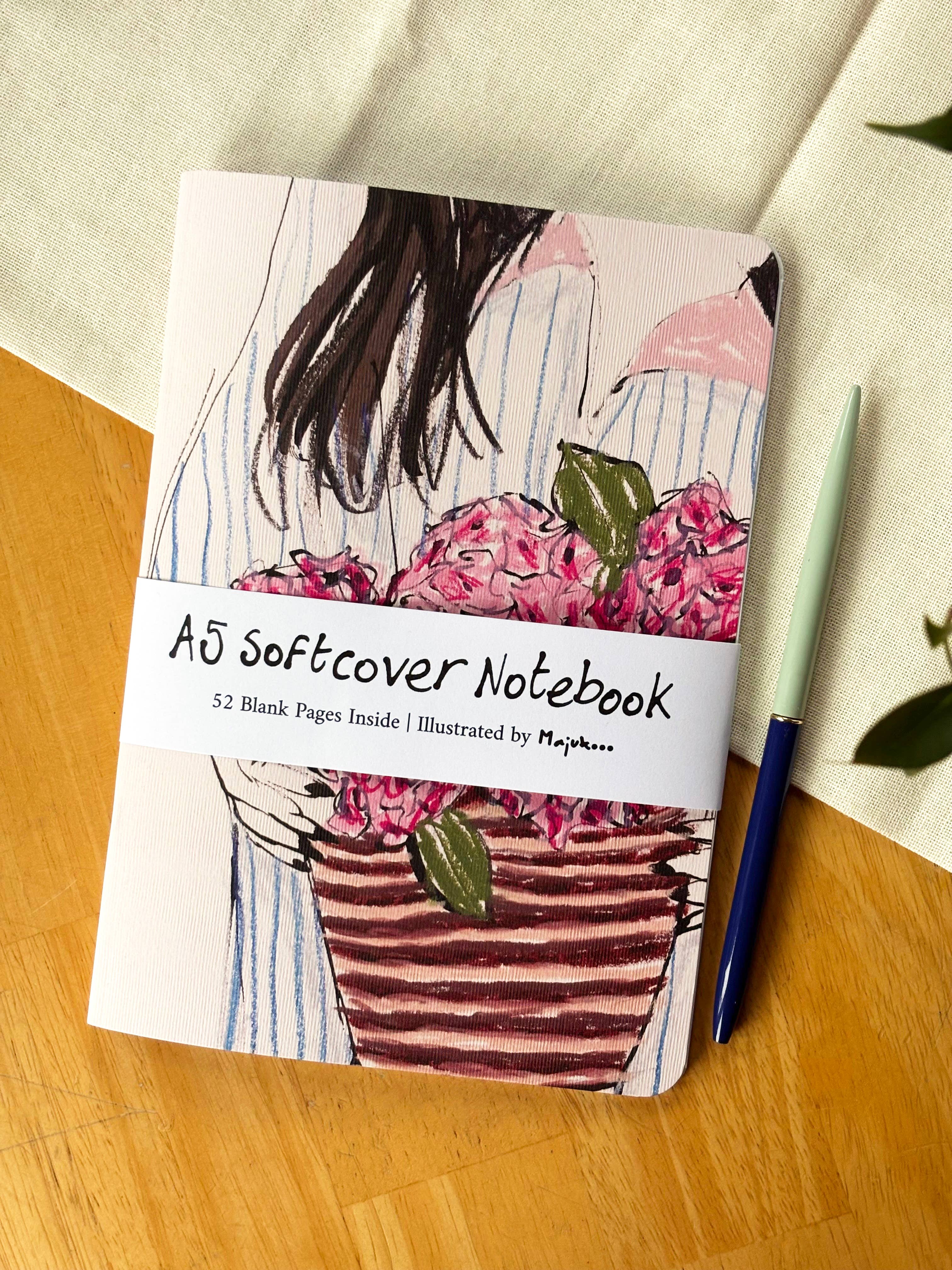 An A5 softcover notebook with an illustrated cover of a woman holding pink flowers, placed on a wooden surface next to a blue pen. A white paper band across the middle reads 'A5 Softcover Notebook' and '52 Blank Pages Inside'.