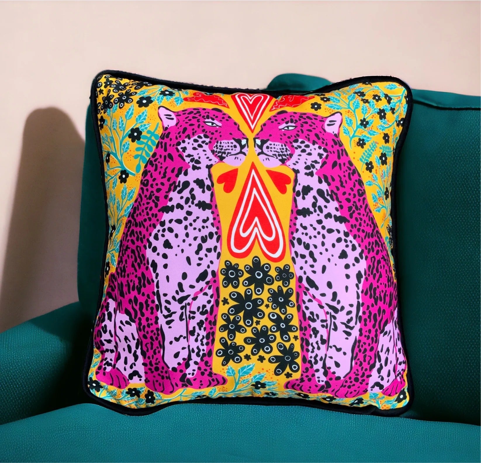 Colourful cushion featuring two pink panthers, red love hearts and green and black flowers against a mustard background, on a sofa.