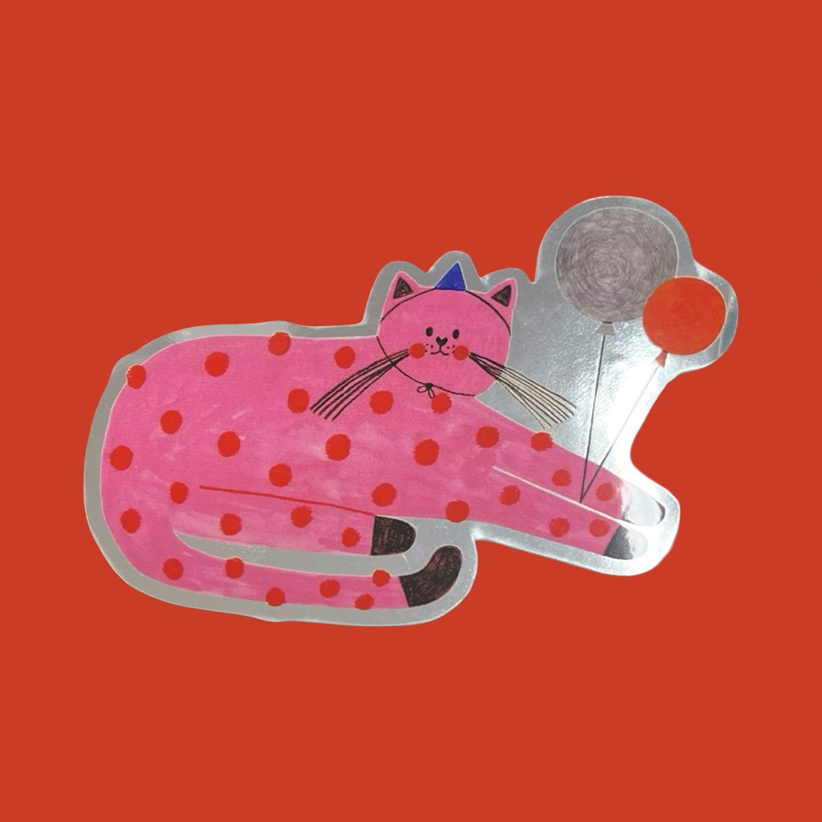 Pink polka dot cat-shaped sticker with a blue party hat on an orange background.