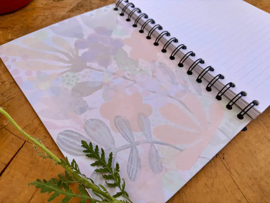 A spiral-bound notebook with a floral pattern on the cover, placed on a wooden surface with a green sprig in the foreground.