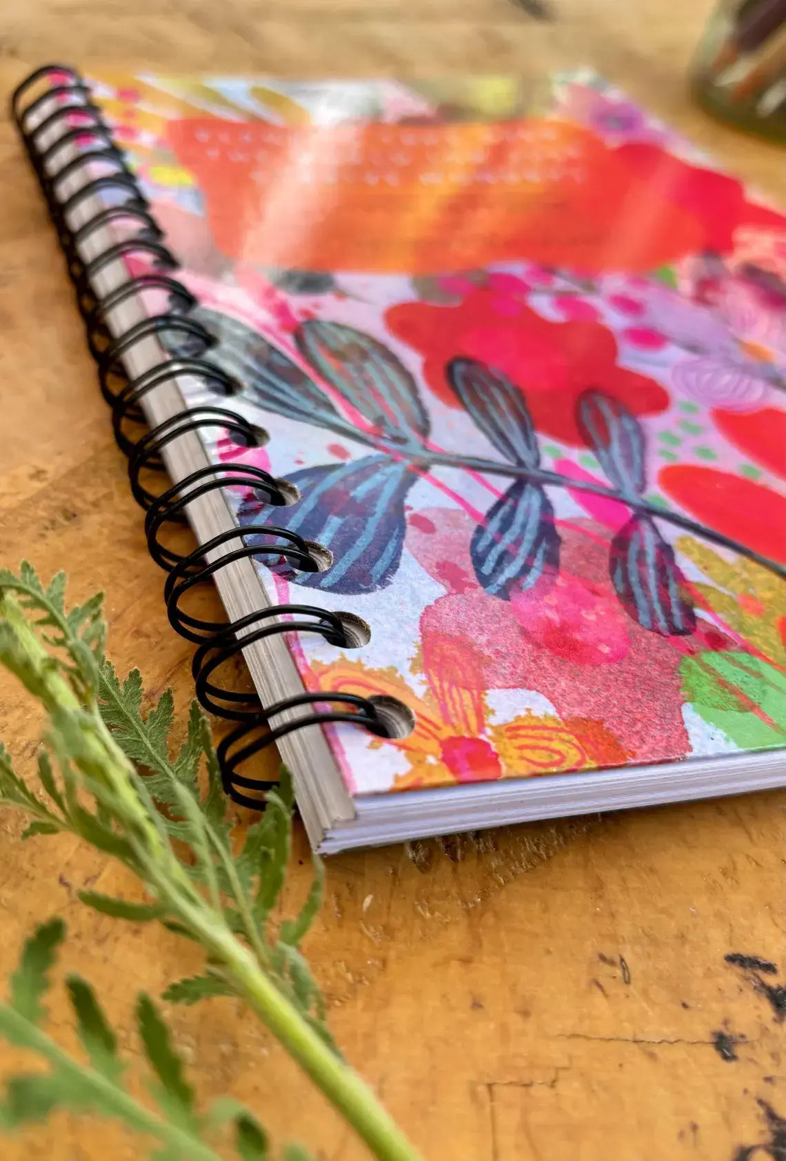 A spiral-bound notebook with a colourful floral cover design, resting on a wooden surface with a green sprig in the foreground.
