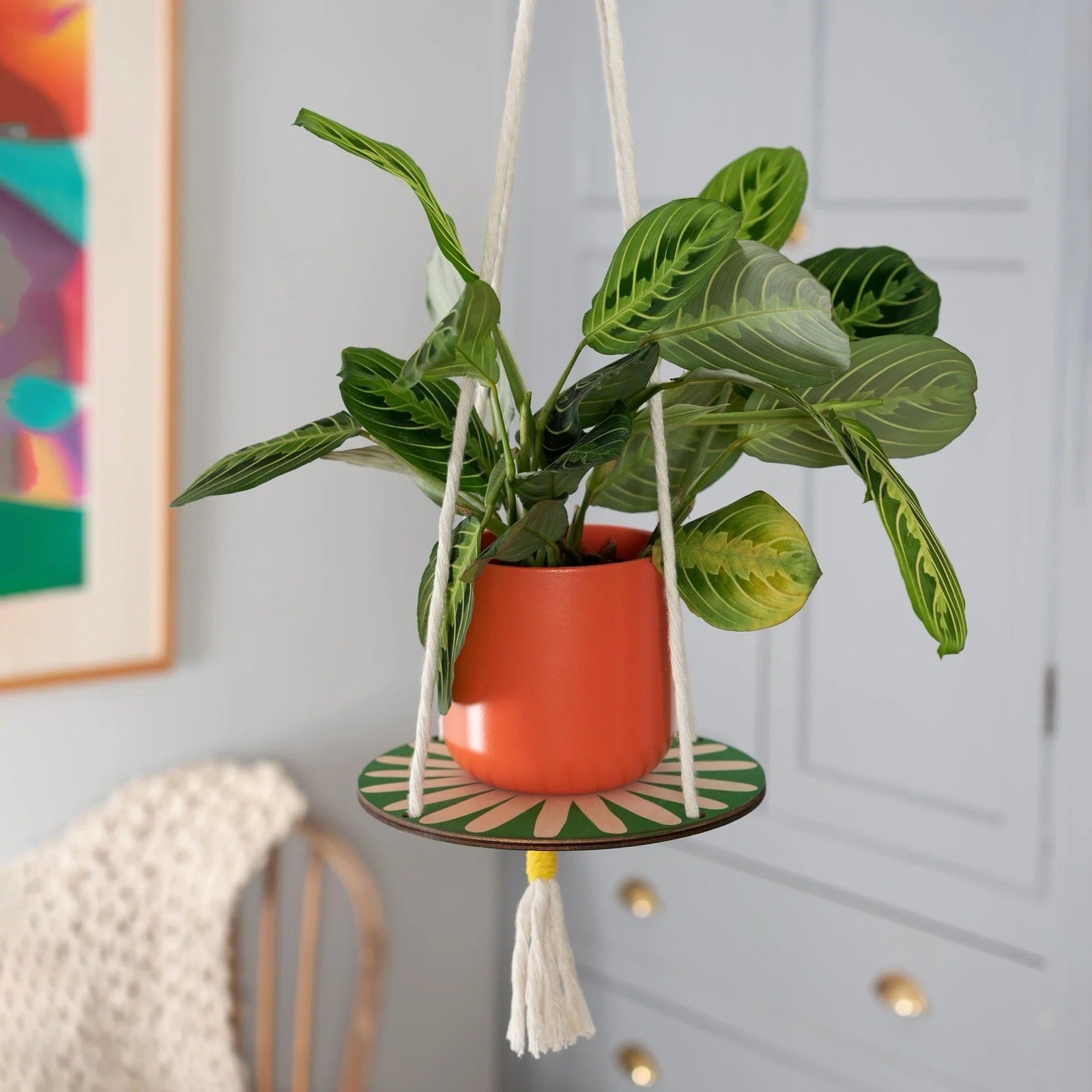 Hanging plant with orange pot on a decorative stand against a light grey wall.