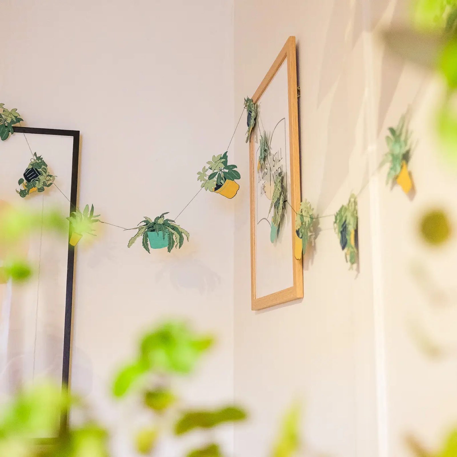 Plant pot garland featuring ten screen-printed house plants in shades of green and mustard hanging on a cream wall.
