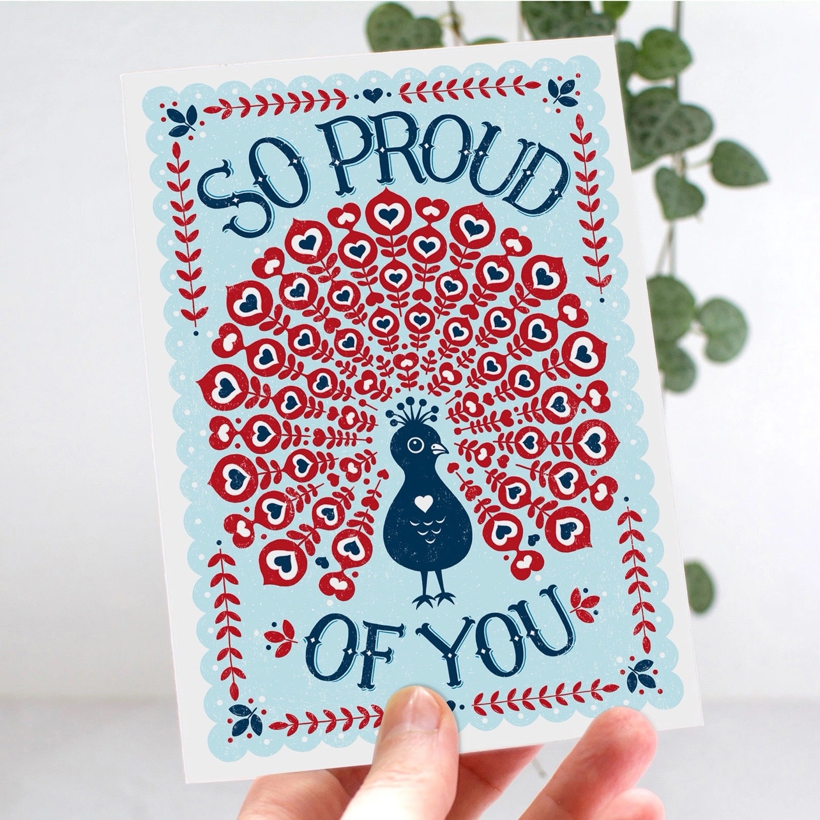 Greeting card with a peacock design and 'So Proud of You' text, held against a light background.