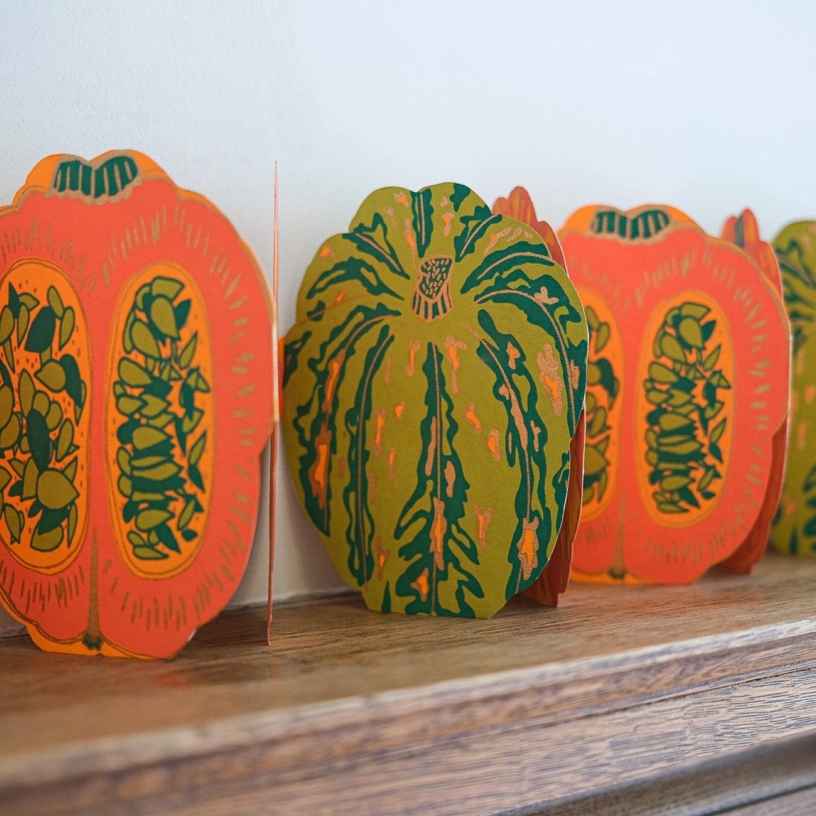 An orange and green pumpkin concertina garland on recycled paper standing on a wooden shelf.