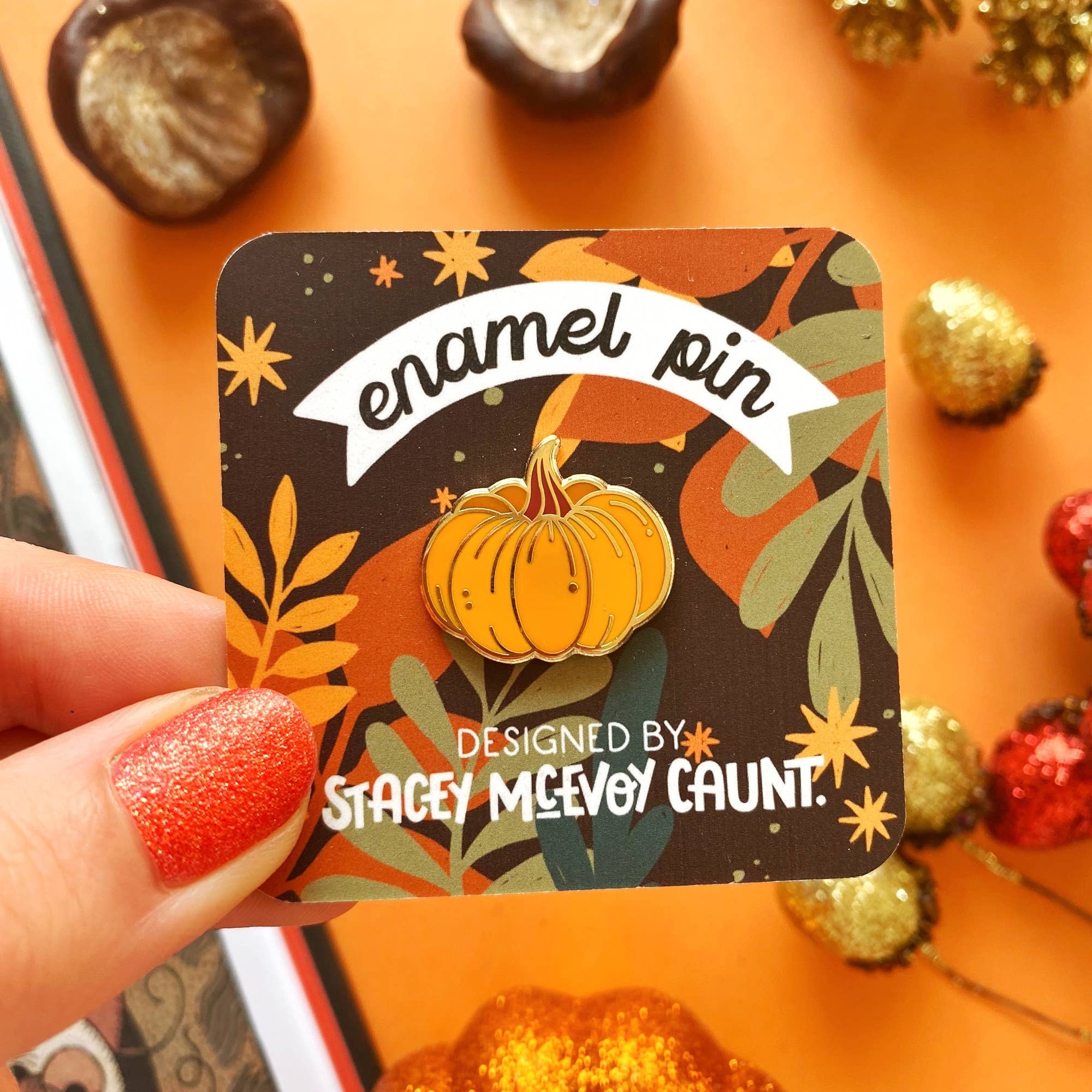 A hand with glittery orange nail polish holds a square display card featuring an enamel pin. The pin is an orange pumpkin on an autumn-themed background with the text 'enamel pin' and 'DESIGNED BY STACEY MEEVOY CAUNT. The backing card is held above an orange table with chestnuts.