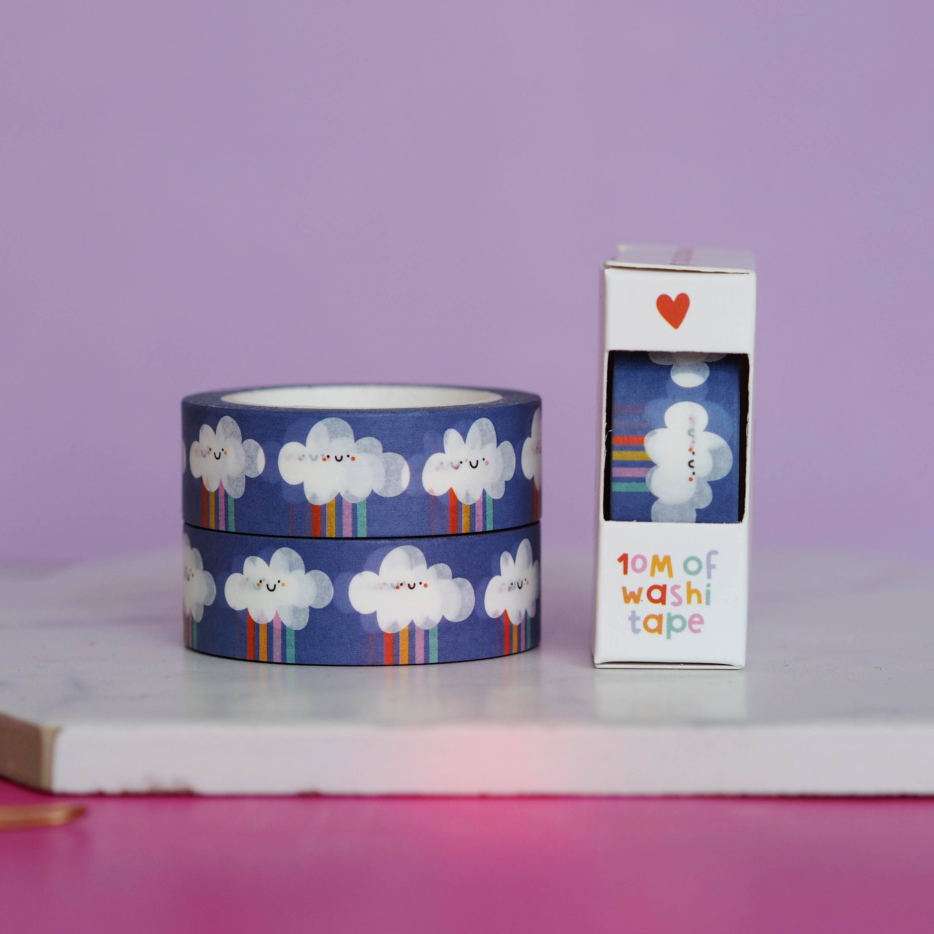 Two rolls of cloud-patterned washi tape with a packaging box on a pink surface and purple background.