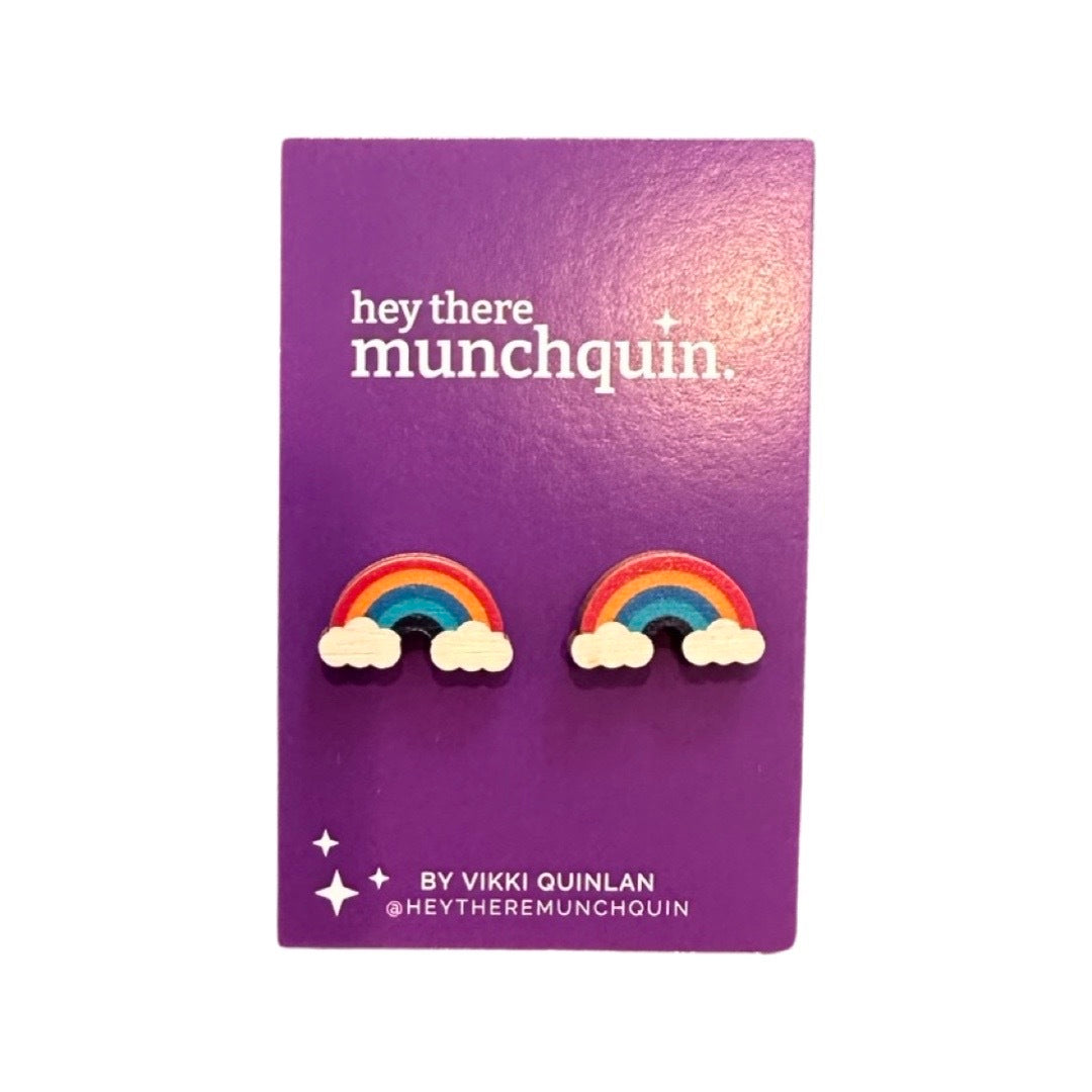 A pair of wooden stud earrings with a rainbow and clouds pattern, displayed on a purple card with 'hey there Munchquin' written on it.