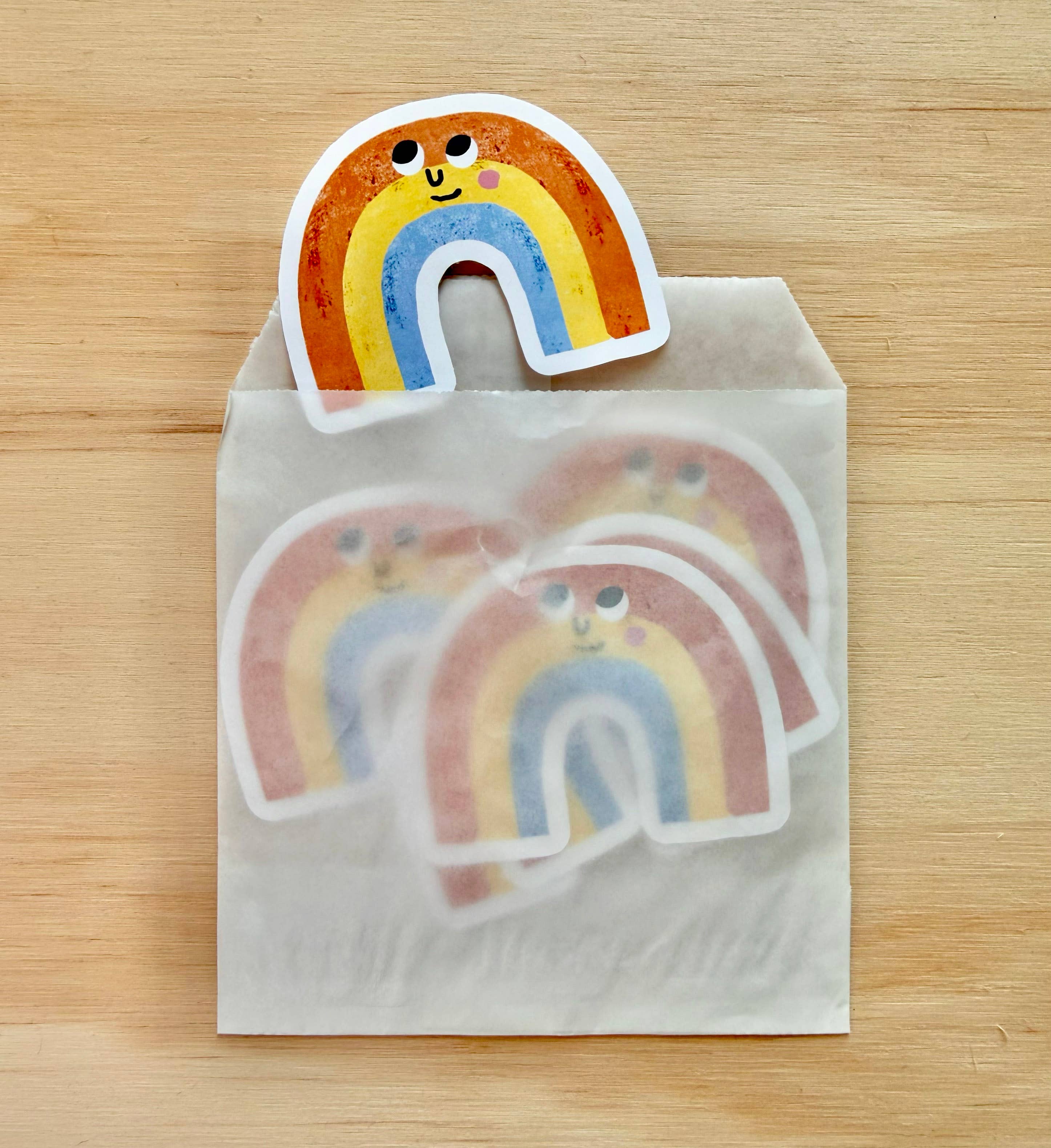 Three die-cut rainbow stickers with a cute cartoon face are displayed on a light wood background. One sticker is shown standing upright on top of a translucent white envelope, while two more are visible inside the envelope.