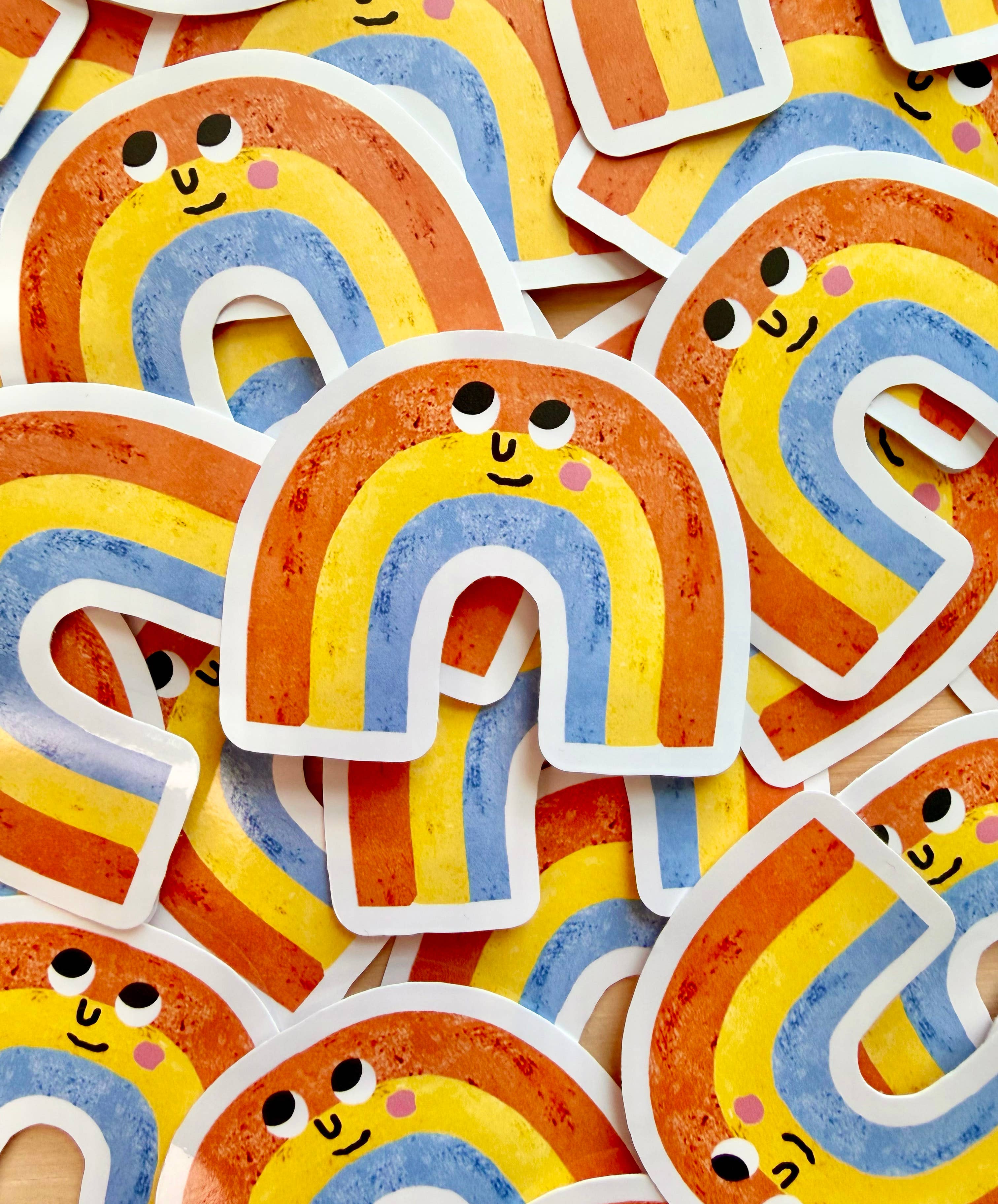 A close-up of multiple die-cut stickers shaped like a rainbow with a colourful rainbow design and a small smiling face, overlapping on a light background.