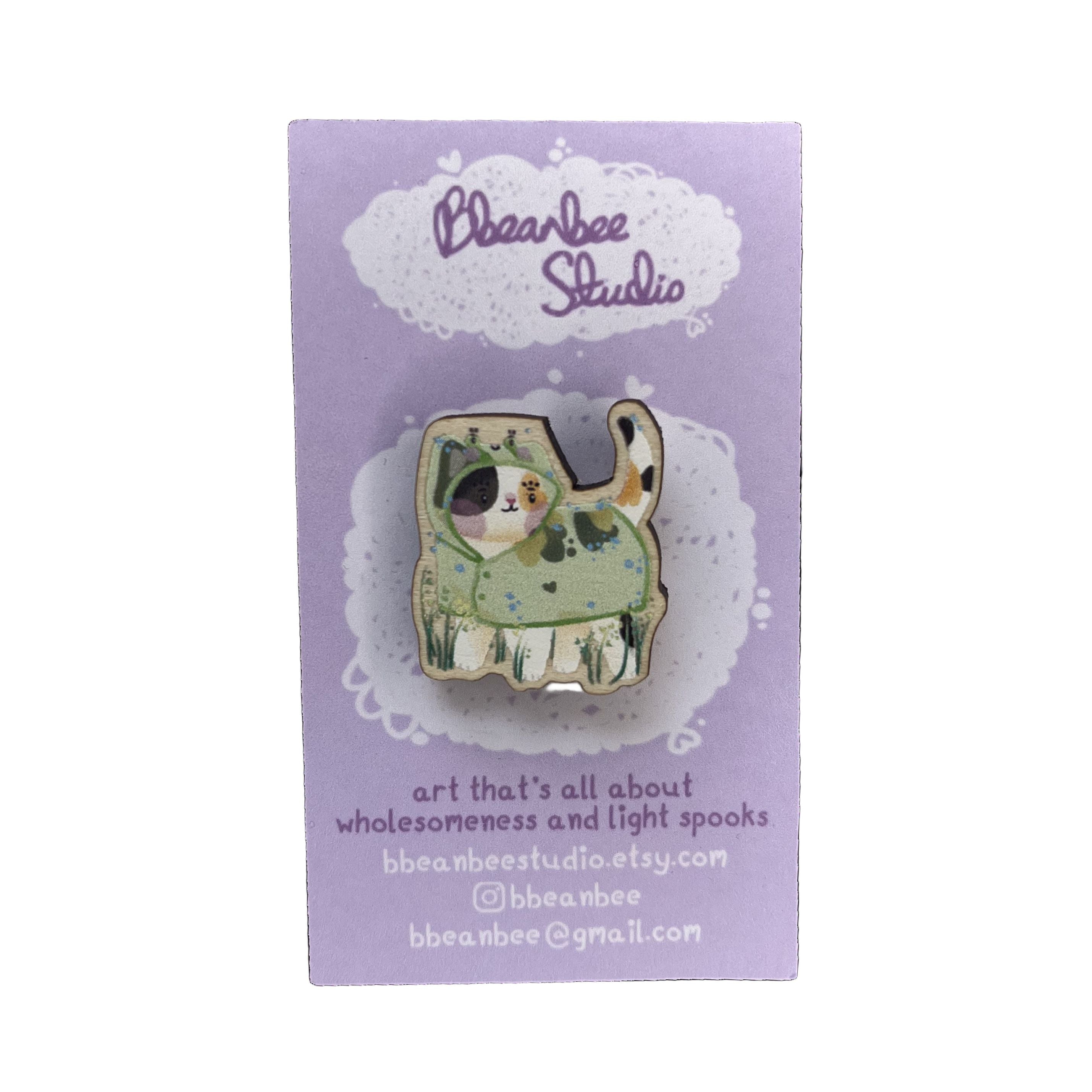Wooden pin badge of a calico cat wearing a frog raincoat, on a purple backing card