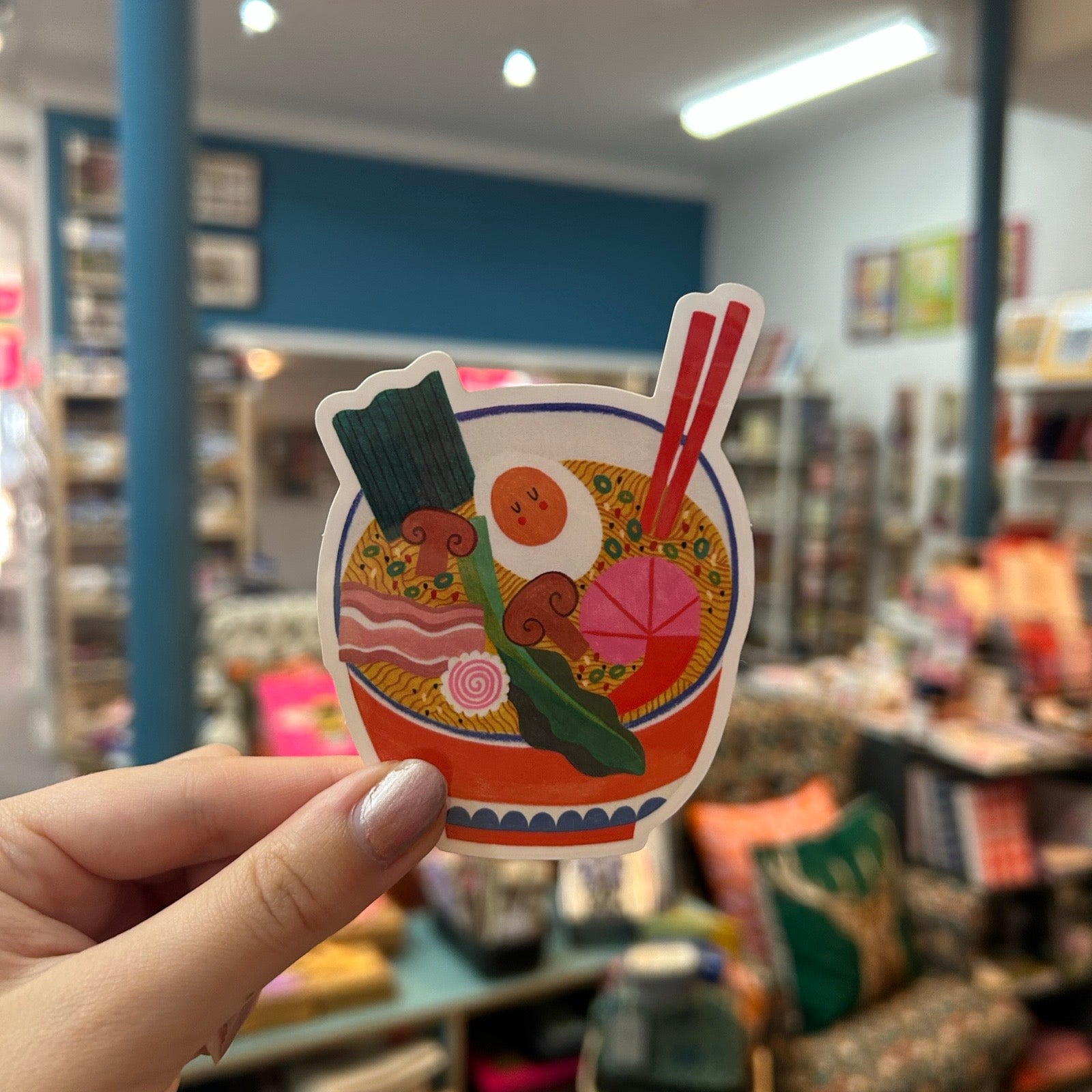 A hand holds a sticker featuring a colourful illustrated ramen bowl with chopsticks, an egg, greens, and pink prawn, set against a blurred shop interior background.